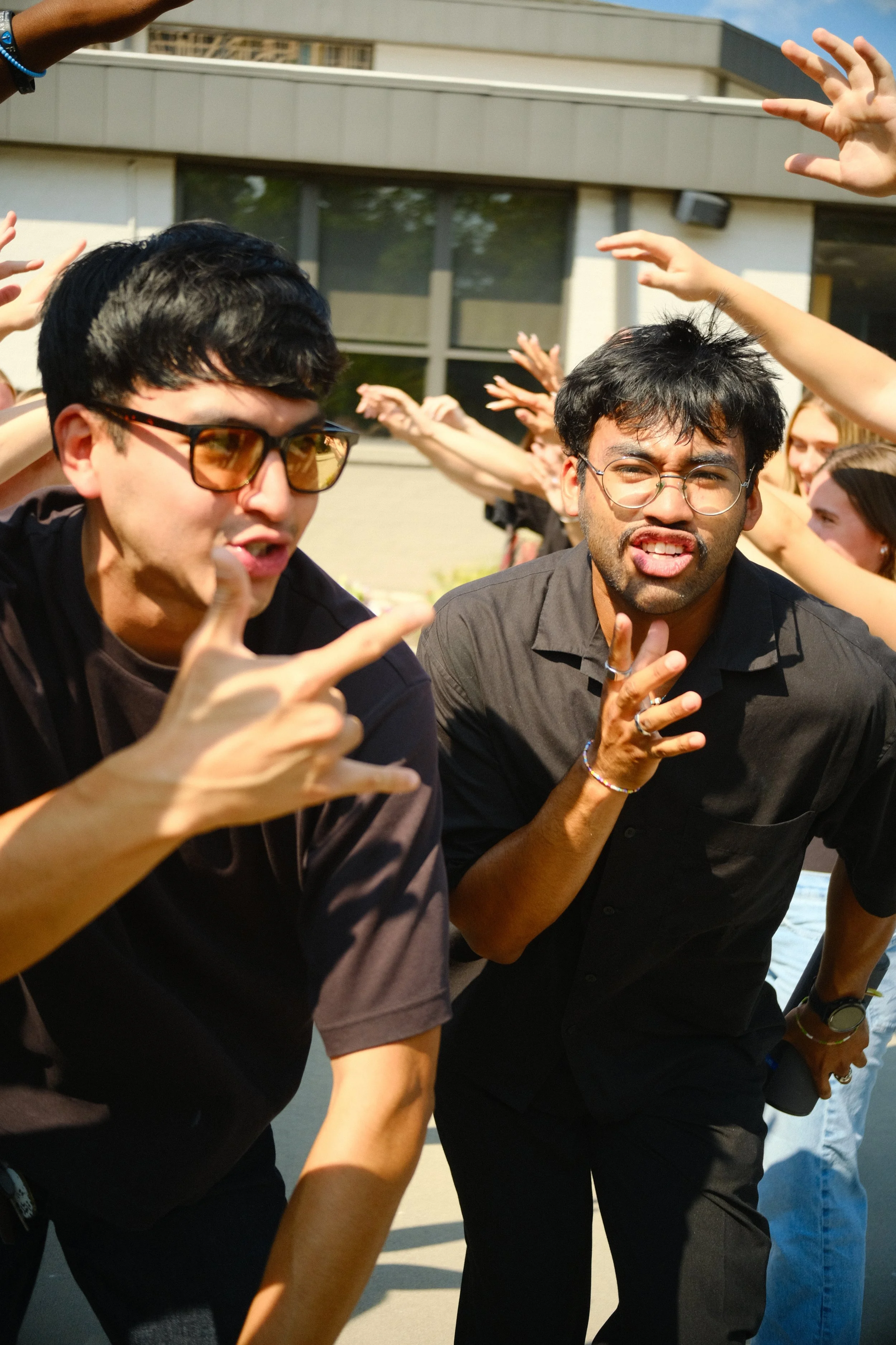 Two men are making faces and gestures with their hands, surrounded by a crowd of people outdoors near a building with large windows.