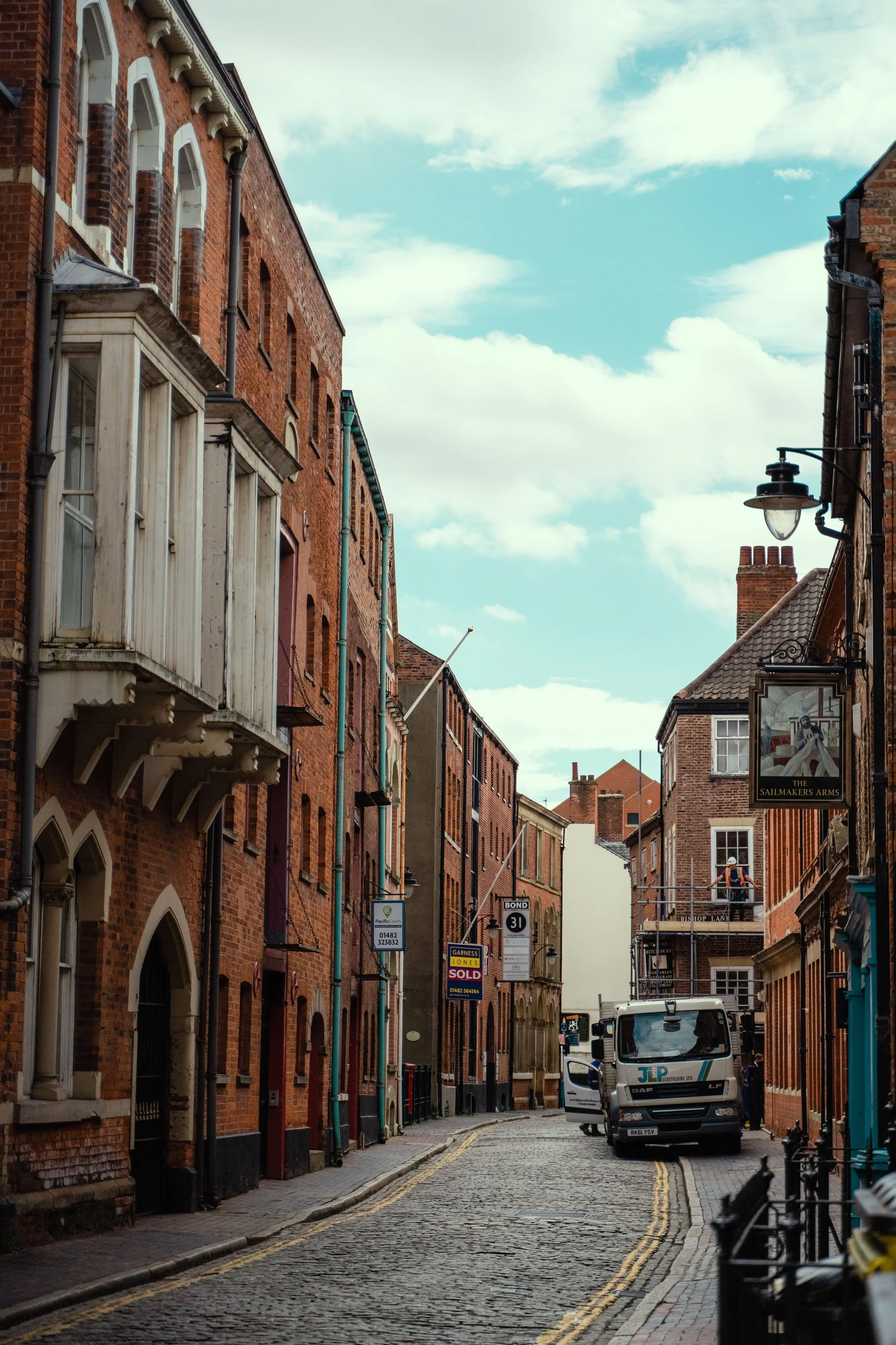 A narrow cobblestone street lined with brick buildings, some with white and wooden balconies, and a few parked vehicles. There are signs on the street, including one indicating a property is sold, and a person with a helmet is visible on a balcony in the background. The sky is partly cloudy.