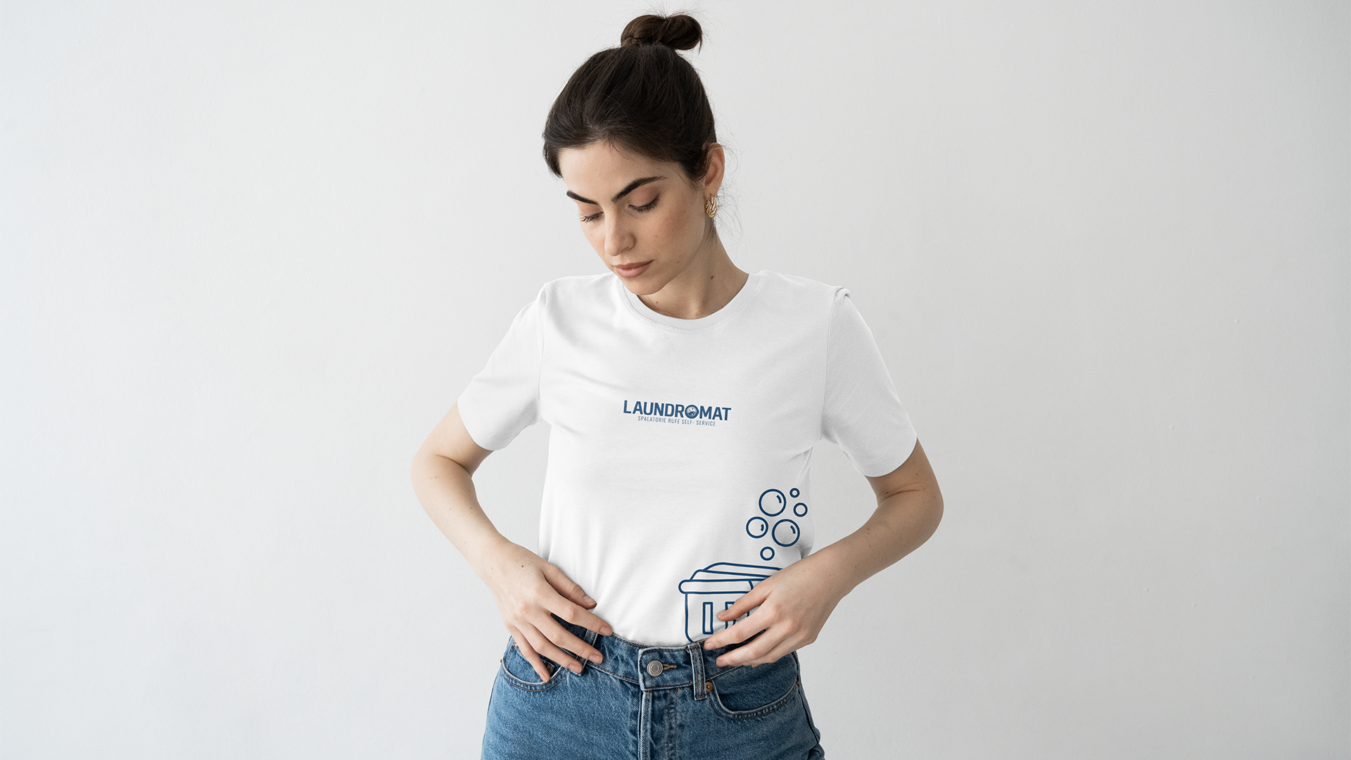 Woman in a white T-shirt with a laundromat logo and cartoon laundry basket graphic, standing against a plain white wall.