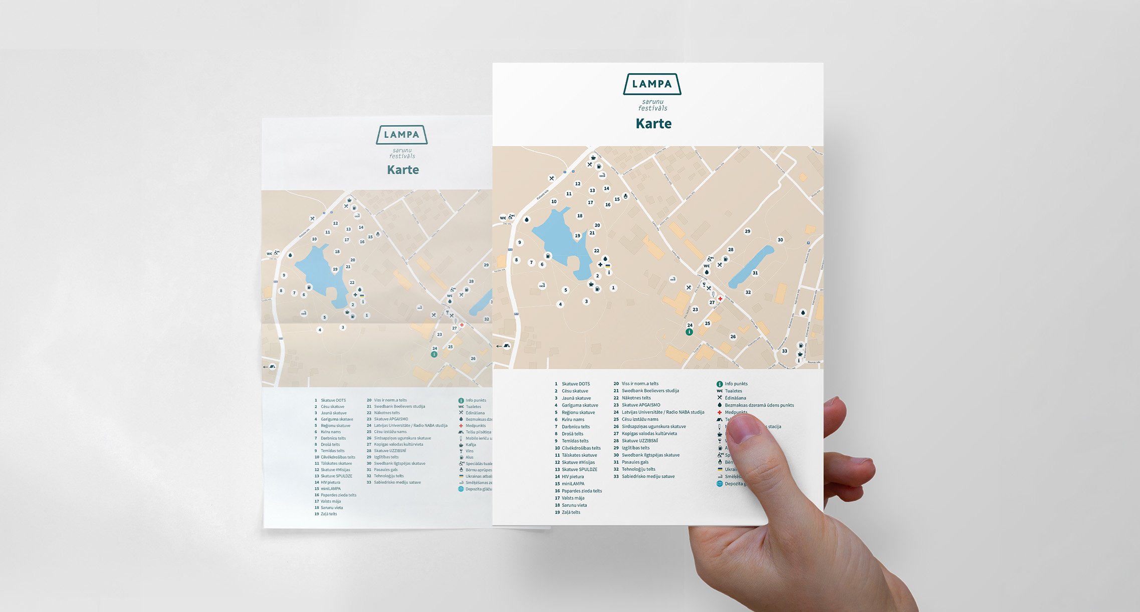 A hand holding a printed map showing locations around a park with lakes, marked with numbered points and symbols, and a legend at the bottom. The map is partially flipped, revealing a second similar map behind it.