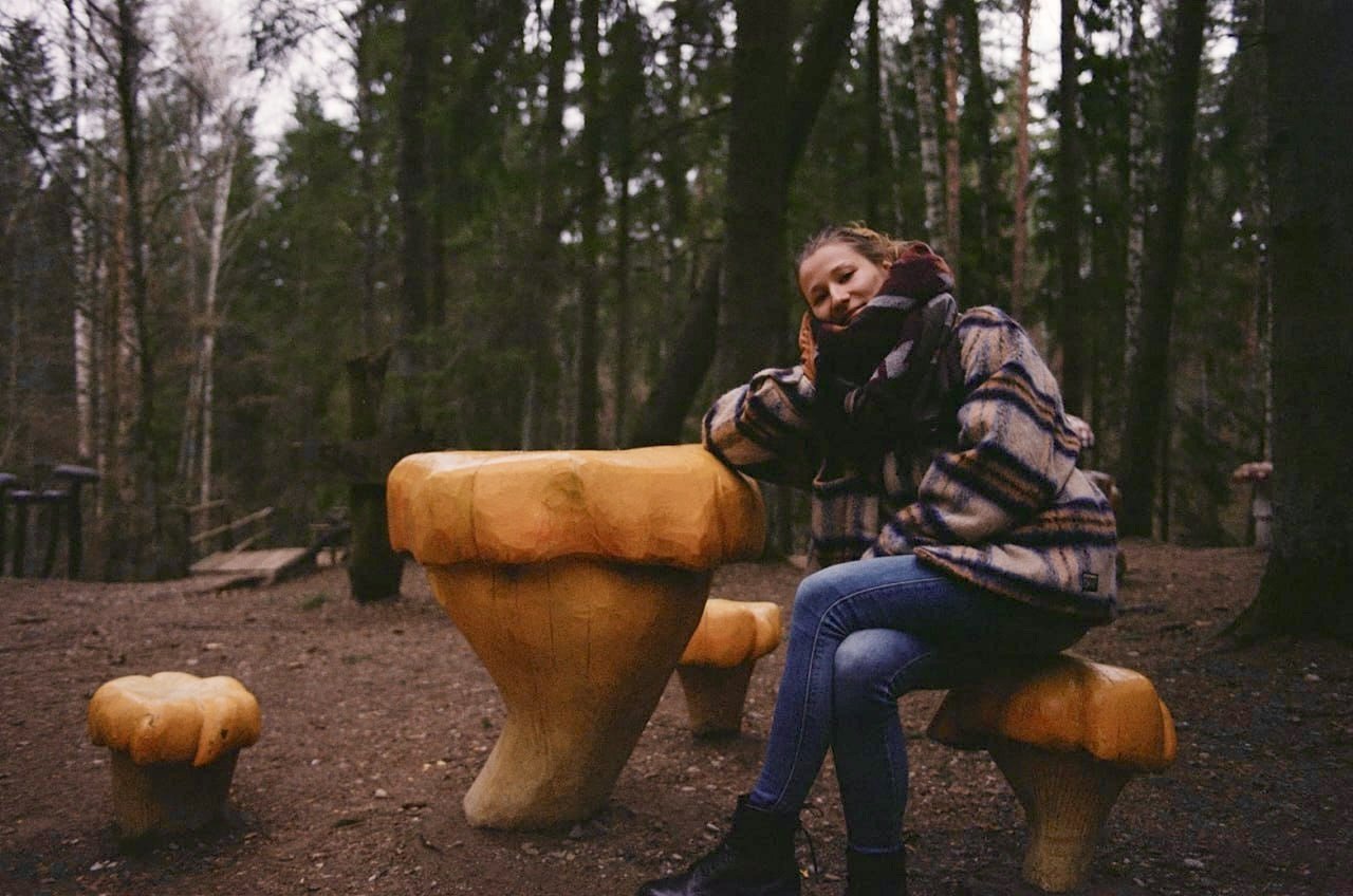 A woman sitting on a wooden bench in a forest, resting her head on her hand and smiling.