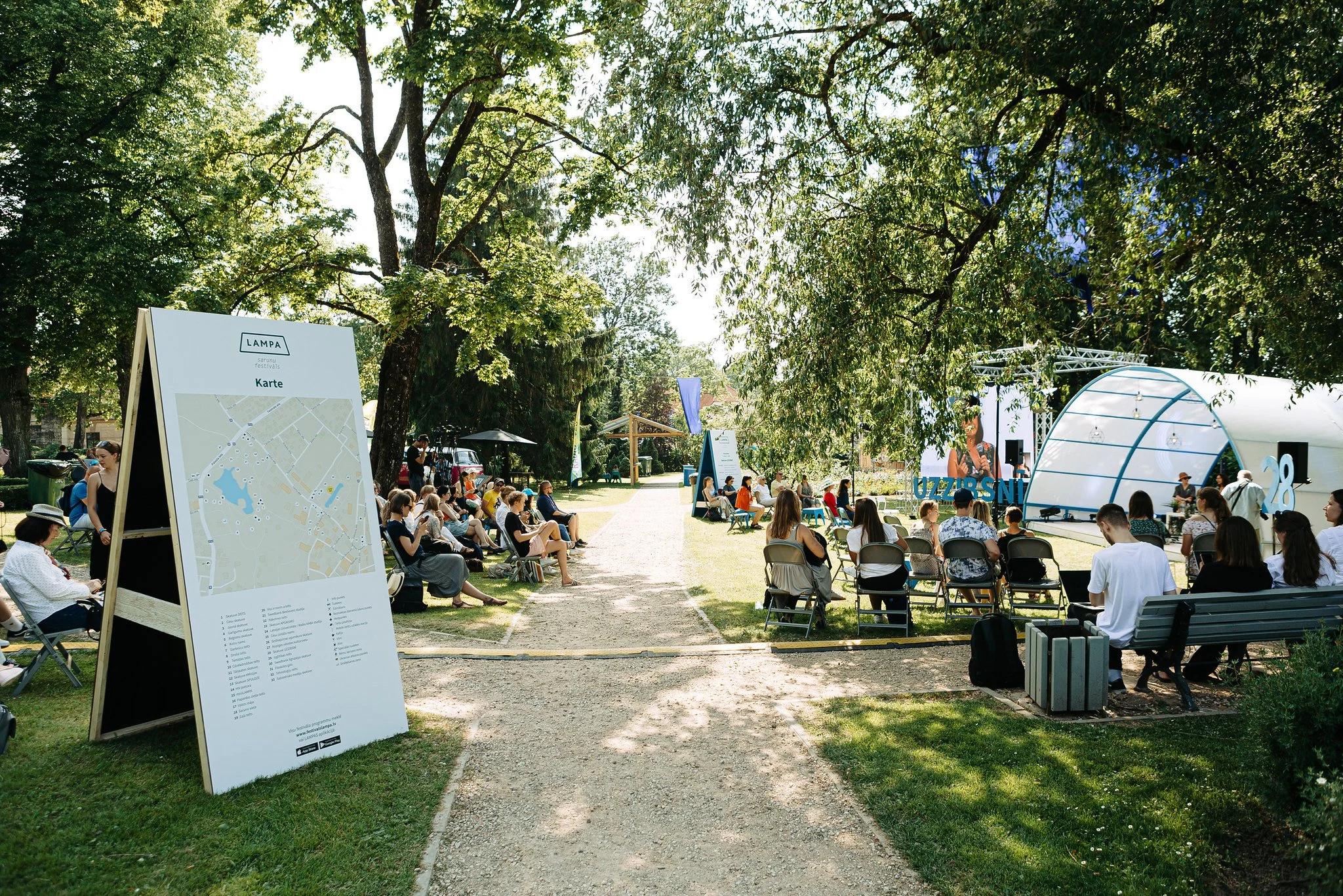 People sitting on chairs outdoors during a sunny day in a park with large trees. There is a large festival map board in the foreground and a stage in the background.