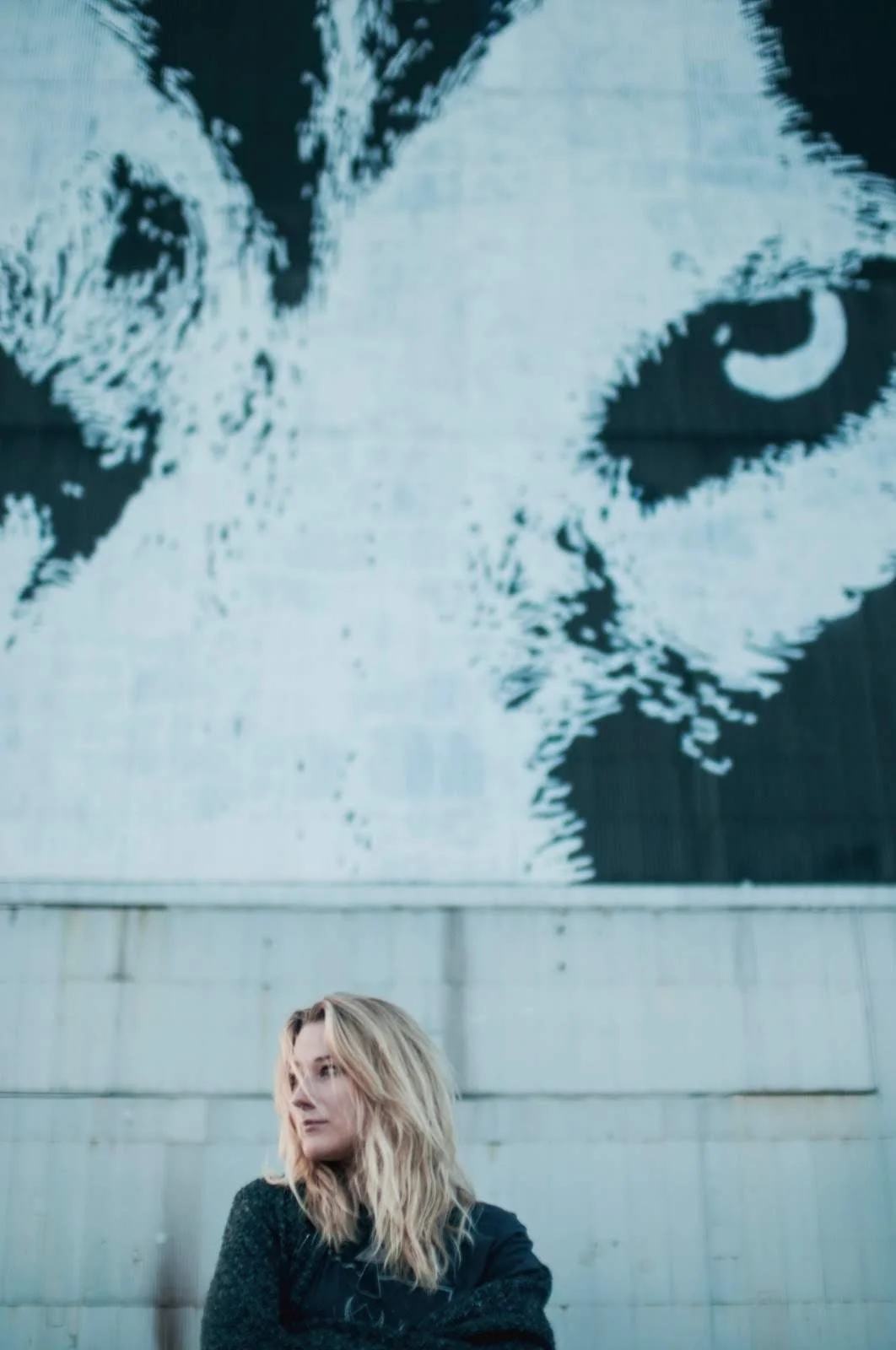 A woman with wavy blonde hair standing in front of a concrete wall beneath a large black and white mural of a cat's face on a building.