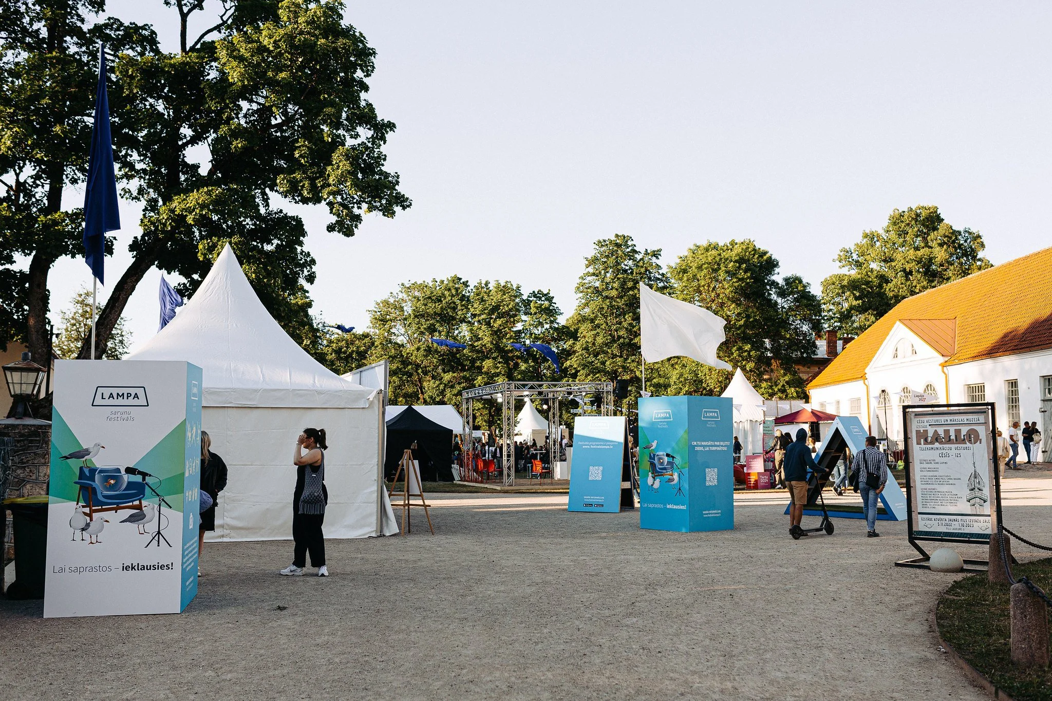 An outdoor festival scene with white tents, informational banners, and people walking around, under a clear sky with trees in the background.