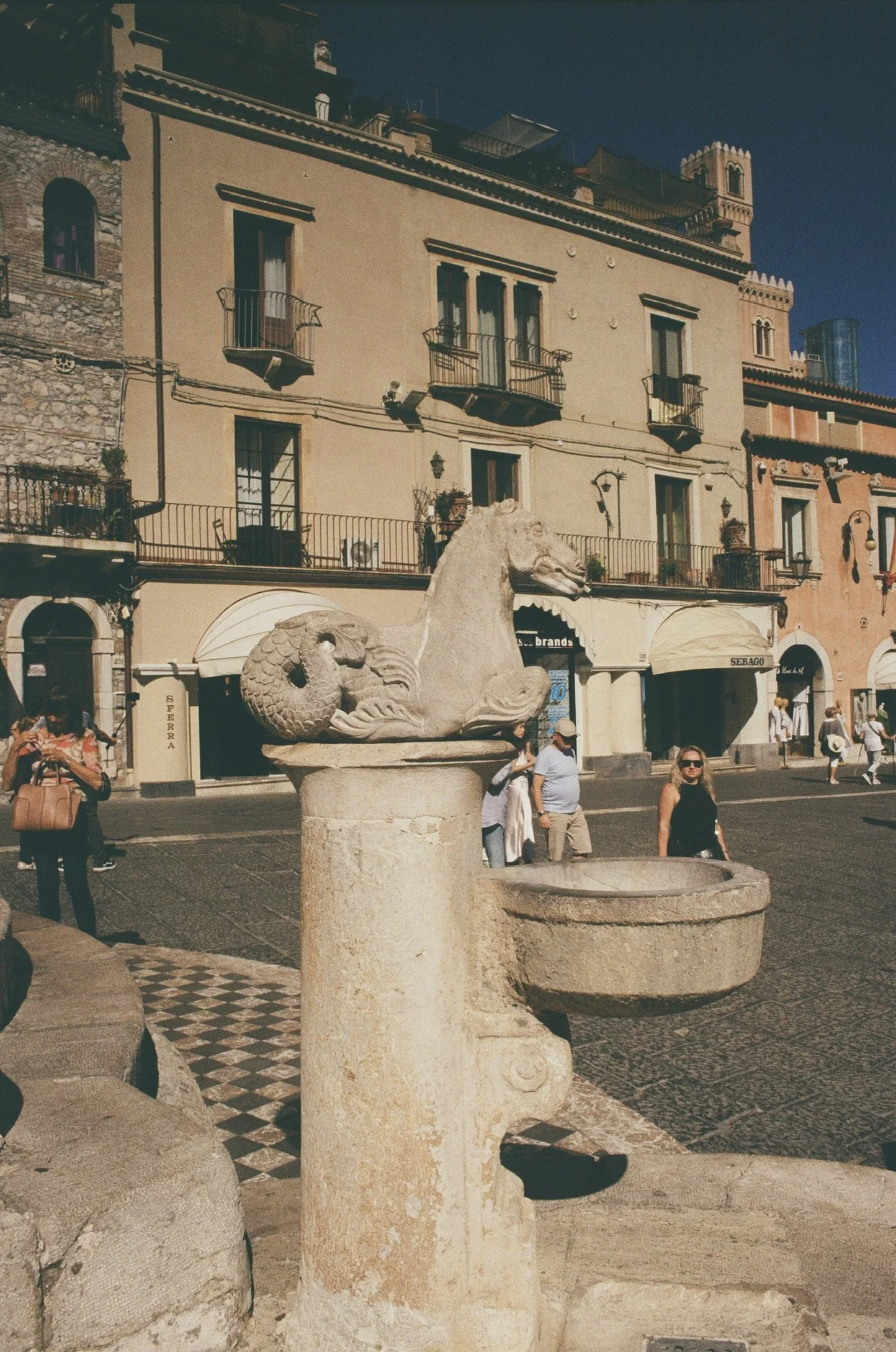 A street scene with a large stone fountain featuring a dragon sculpture in the foreground. Behind it, there are a few people walking and standing. The background shows multistory buildings with balconies, windows, and decorative architecture, suggesting a European city setting.