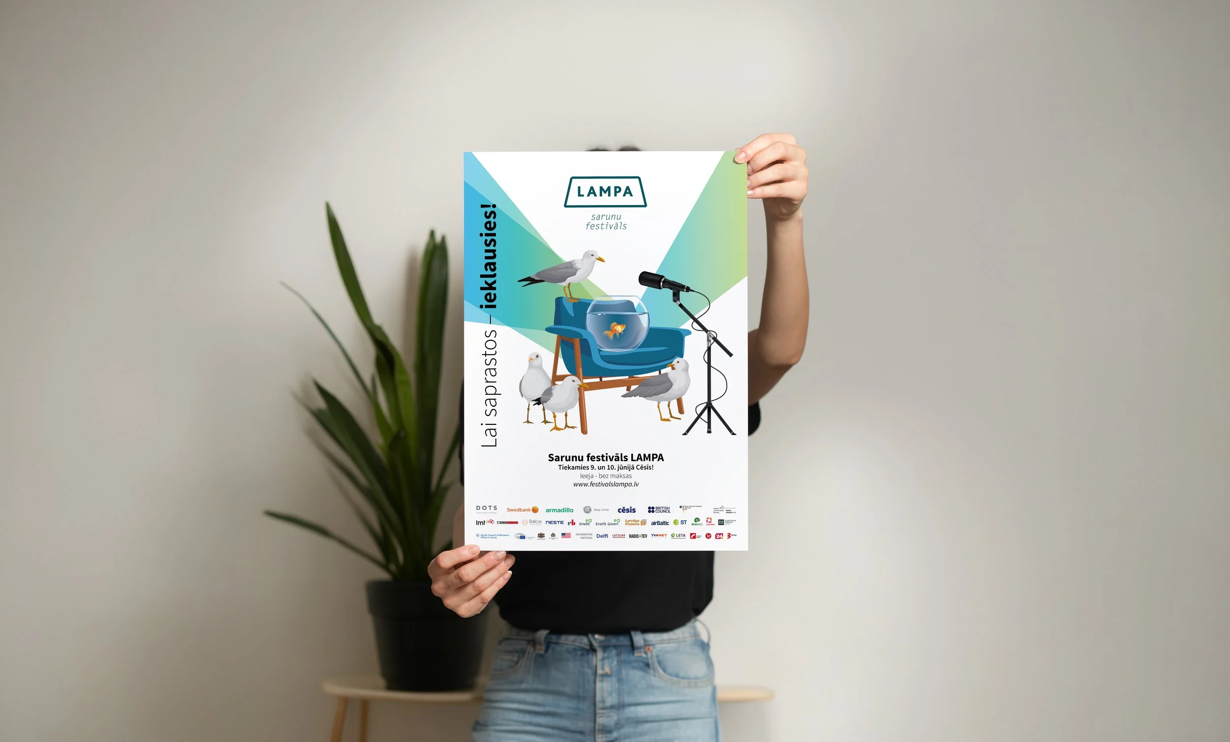 Person holding a colorful flyer for a Seagull Festival with an illustration of seagulls, a chair, and a fishbowl, with a plant in the background.