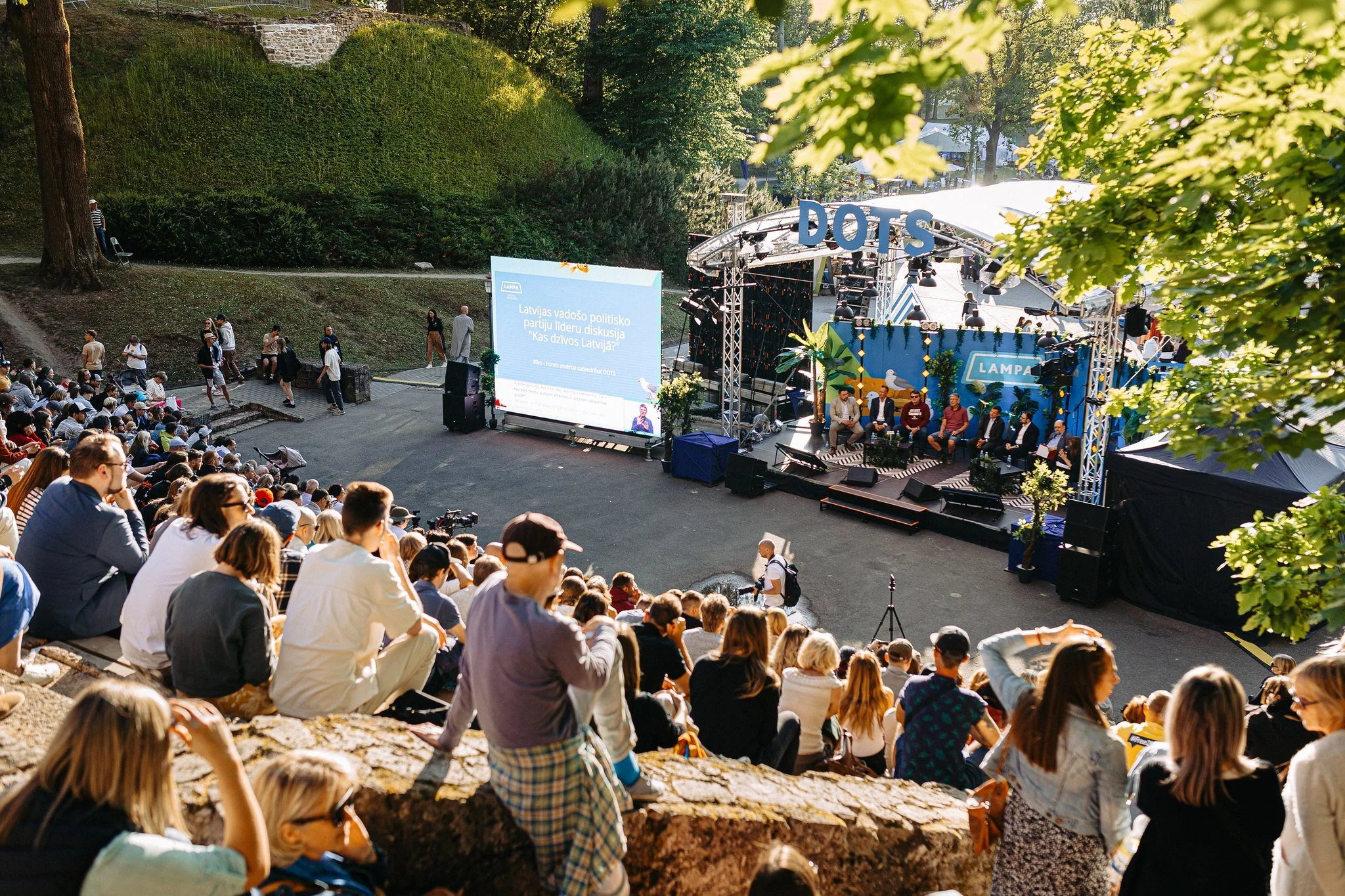 An outdoor event with a seated audience watching a panel discussion on a stage labeled 'DOTS,' set in a park with green trees and a grassy hillside.