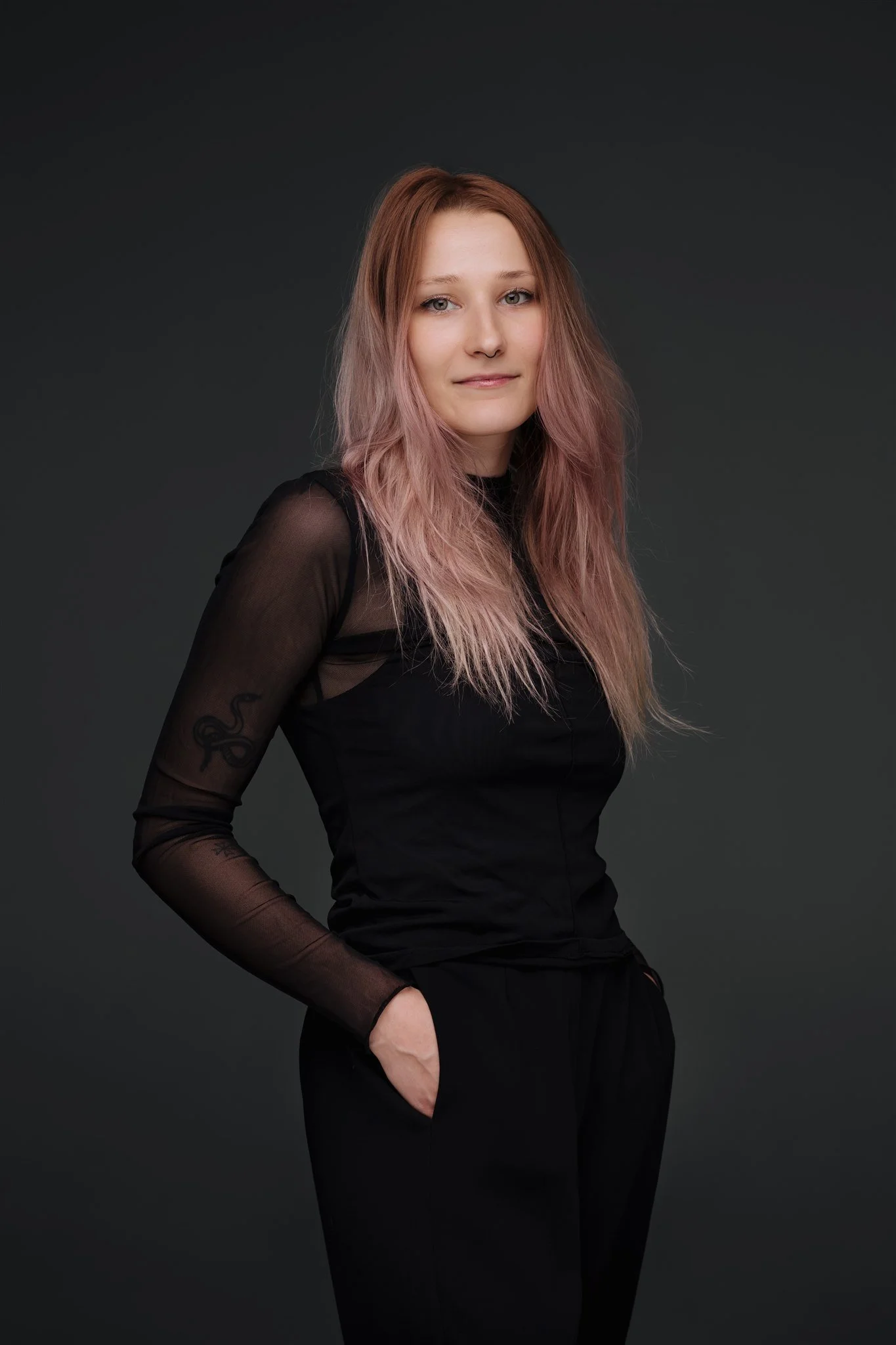 A young woman with long, wavy pink hair and blue eyes, wearing a black top with sheer long sleeves, standing against a plain dark background.