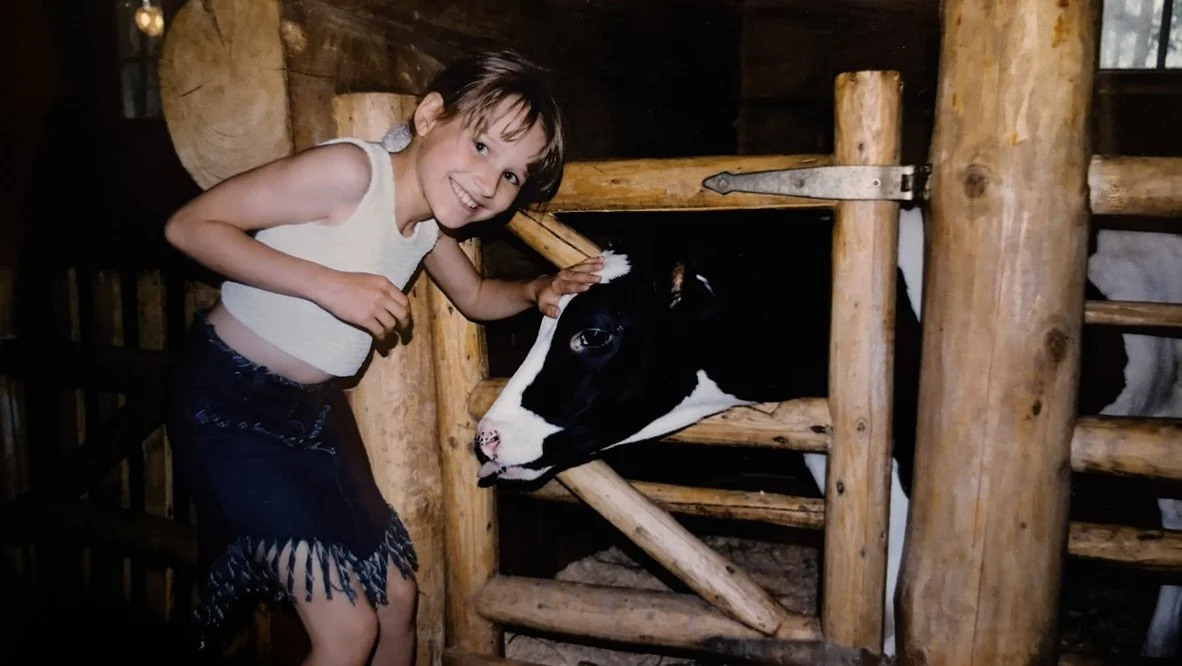 A young girl with a short haircut and wearing a white sleeveless top and a fringed skirt is smiling and petting a black and white calf through a wooden fence in a rustic barn setting.