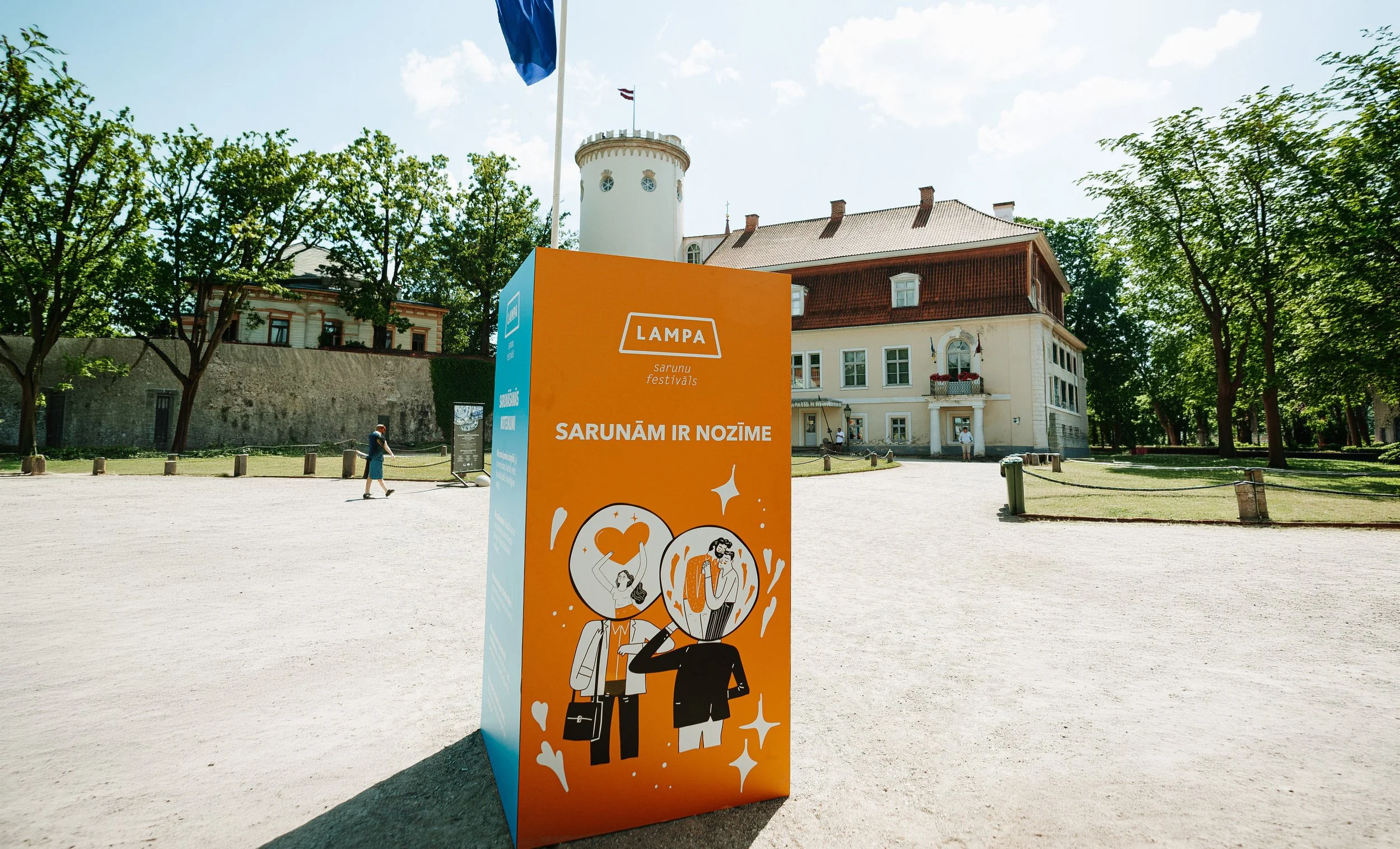 A bright orange festival sign with illustrations of people celebrating, set outdoors in front of historic buildings and lush green trees on a sunny day.