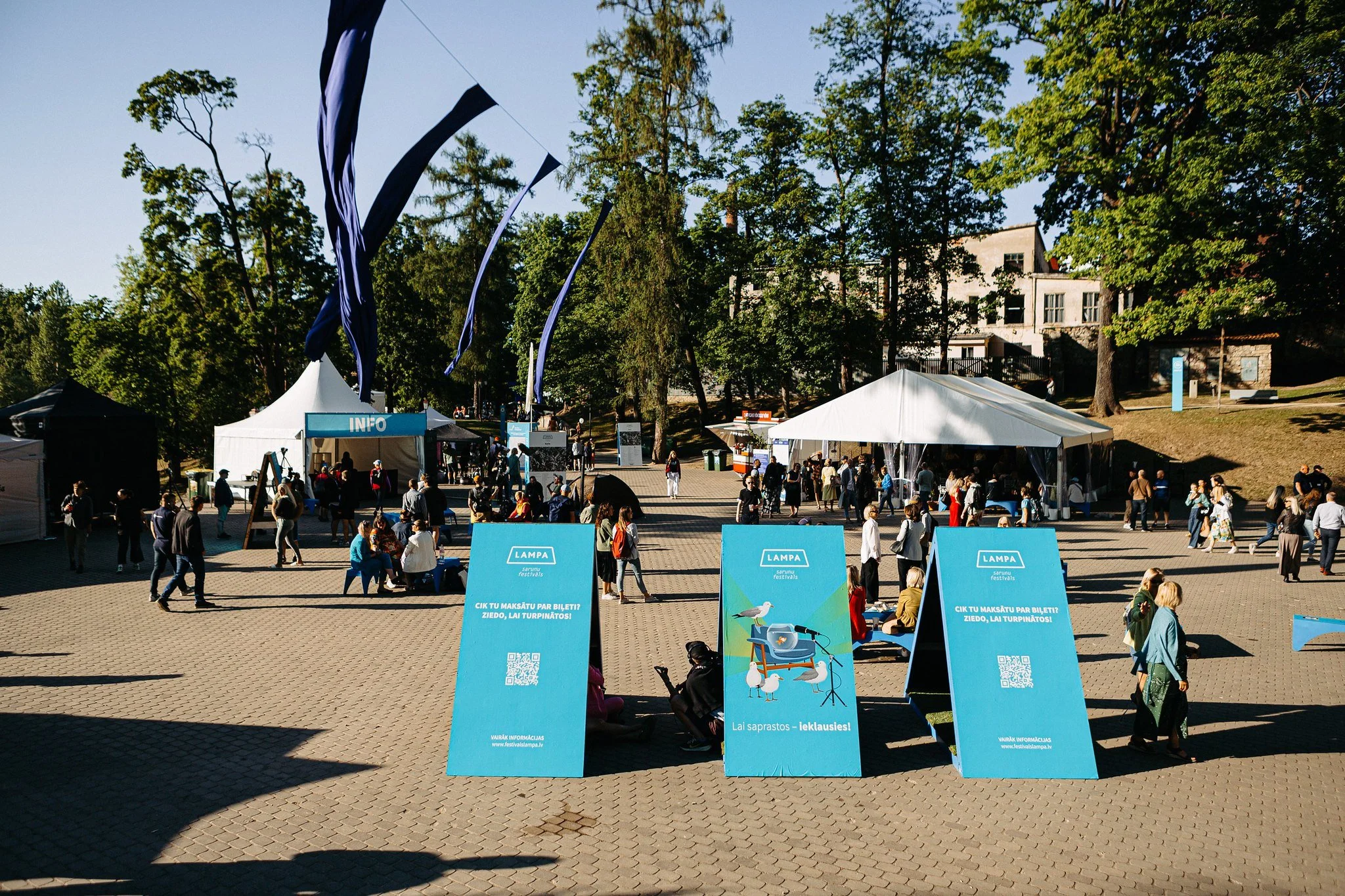 An outdoor festival scene with people walking and sitting under tents, with banners and signs in Latvian, trees, and a building in the background.