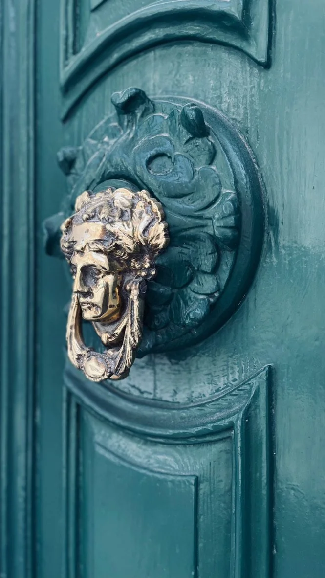A close-up of a green door with a decorative lion head door knocker and a metal ring in its mouth.