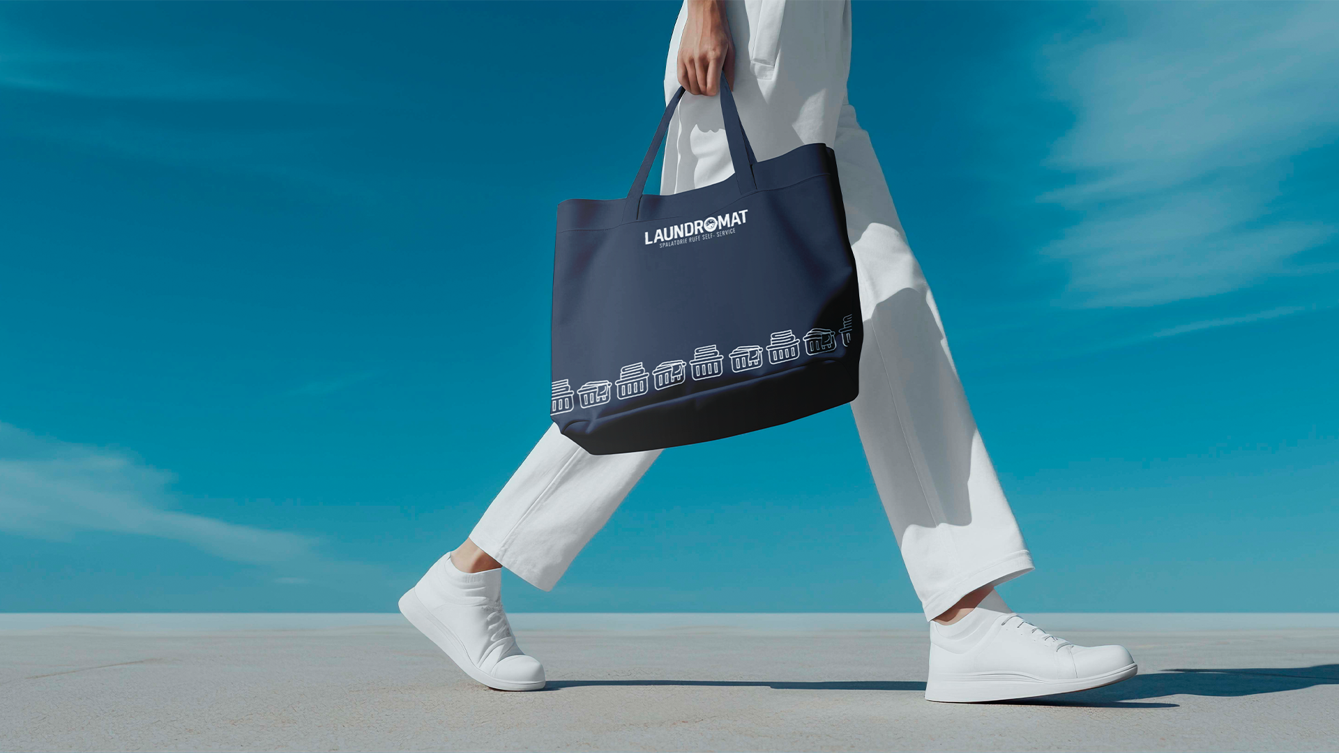 Person wearing white sneakers and white pants, carrying a navy blue tote bag with the word 'LAUNDROMAT' and graphic icons of laundry baskets, against a bright blue sky.