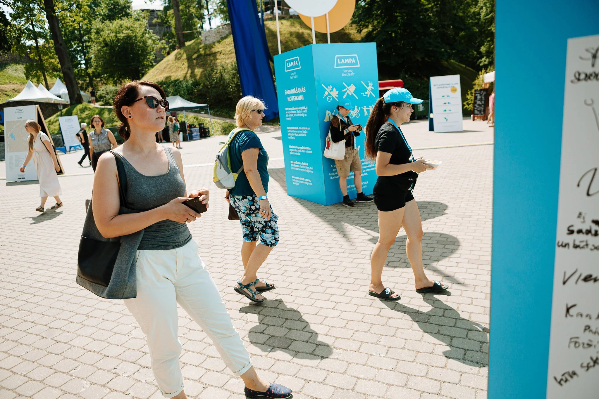 People walking outdoors at a festival with informational signs and banners. Sunny day with trees and tents in background.