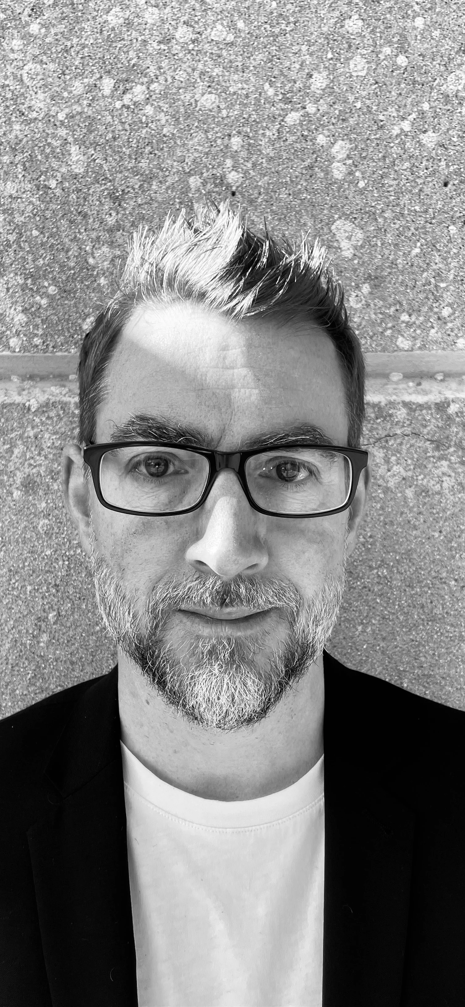 A black-and-white close-up selfie of a man with glasses, a beard, and styled hair, standing against a textured wall background.