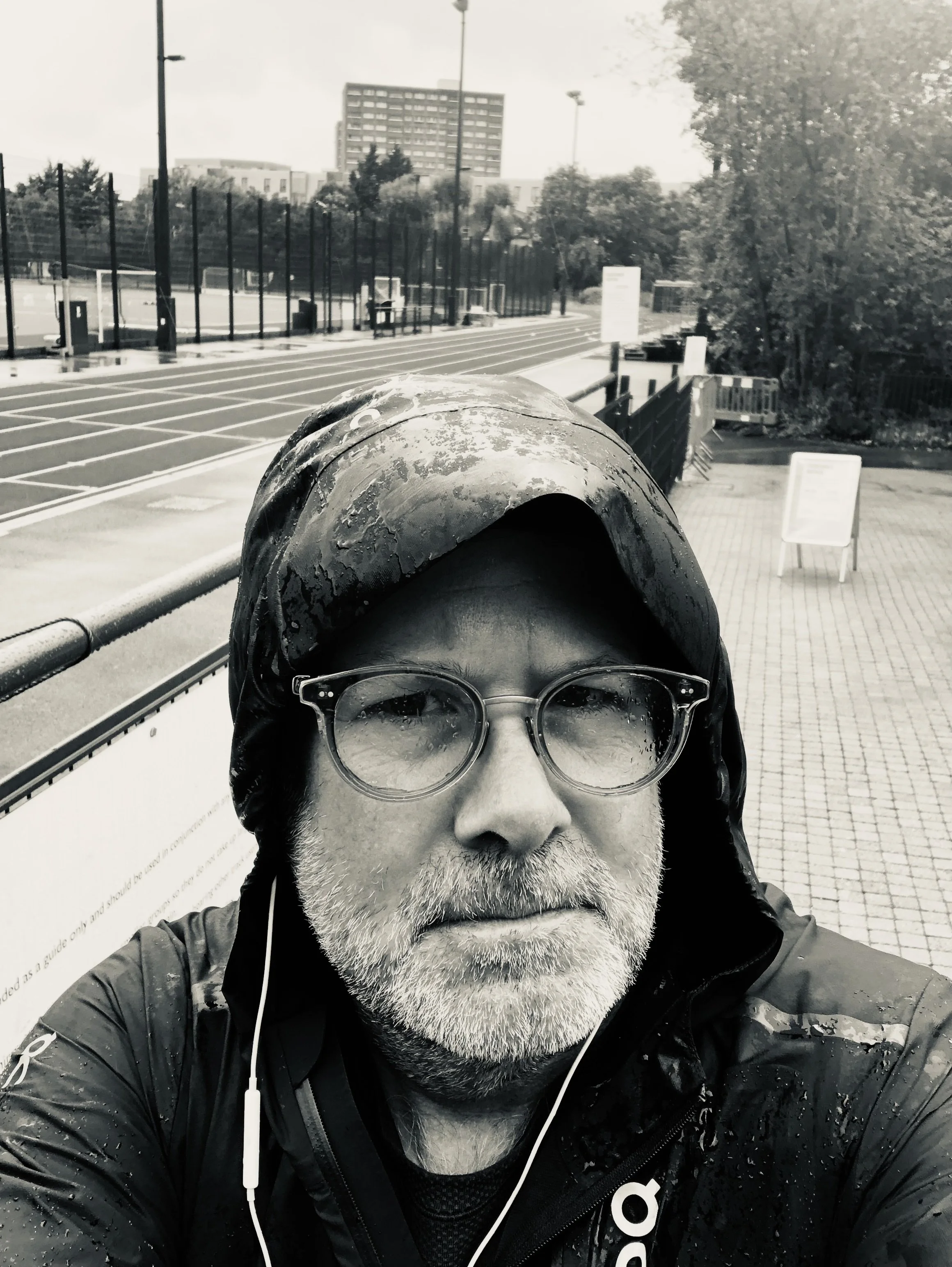 Close-up of a man with glasses, a gray beard, wearing a hooded jacket with raindrops on it, standing outside in a rainy setting, with a park and a large building in the background.