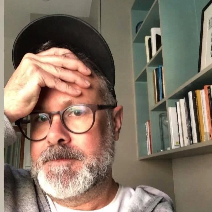 A man with glasses and a gray beard, wearing a black cap, holding his forehead with one hand, in a room with a bookshelf in the background.