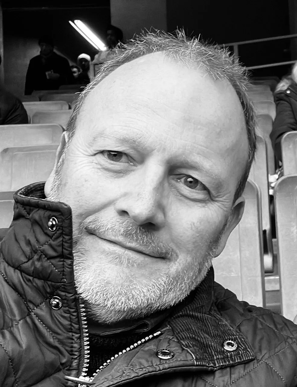 Close-up black and white photograph of a middle-aged man with light facial hair and short hair, smiling slightly, wearing a quilted jacket, in an indoor setting with empty seats and a background of people and lights.