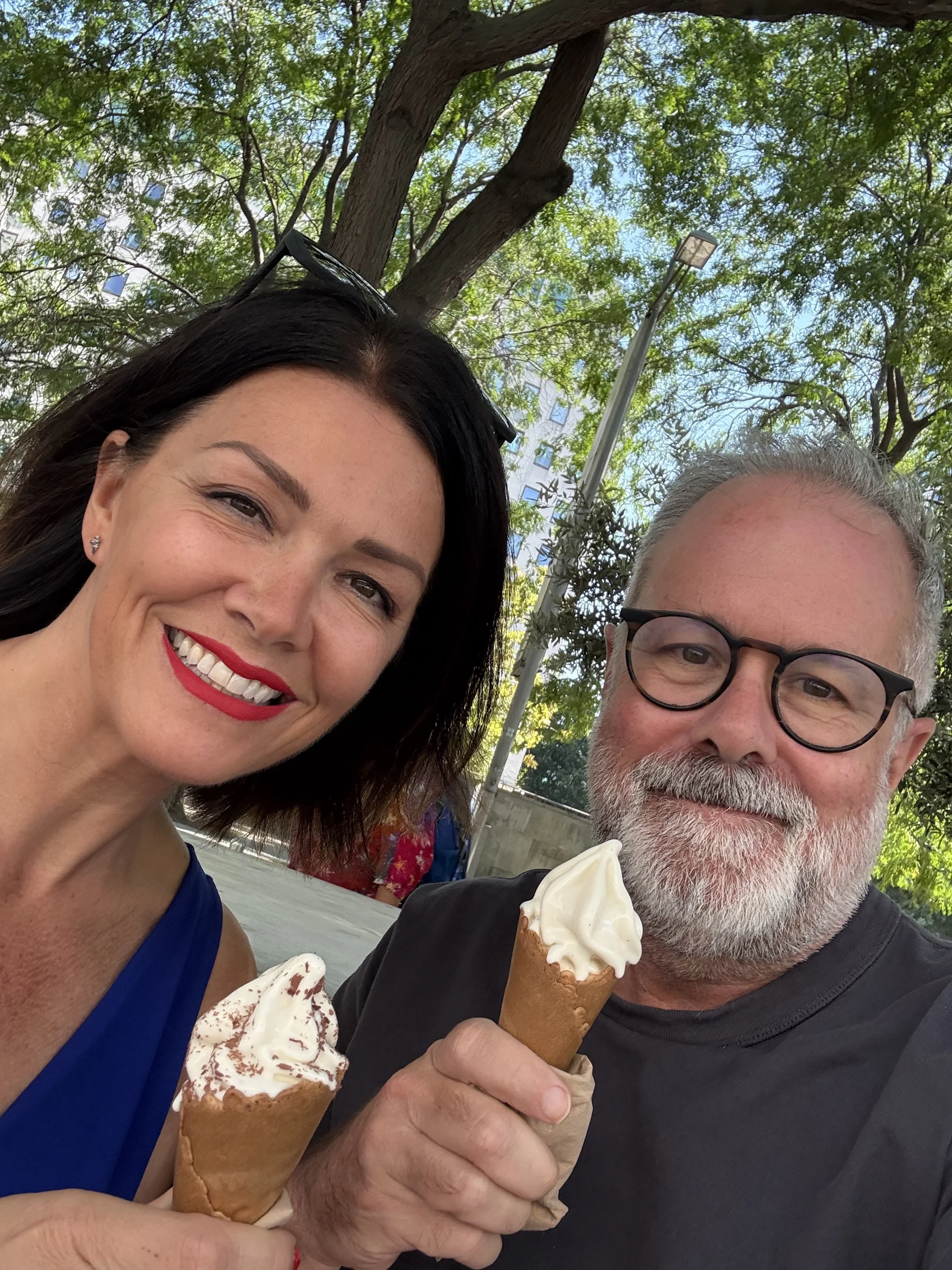A woman and a man enjoying ice cream cones outdoors, sitting at a park or similar area with trees and a lamp post in the background.
