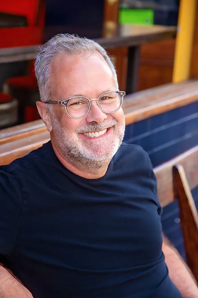 A smiling middle-aged man with gray hair, beard, and glasses, wearing a black t-shirt, sitting outdoors at a restaurant or cafe with wooden benches and colorful background objects.