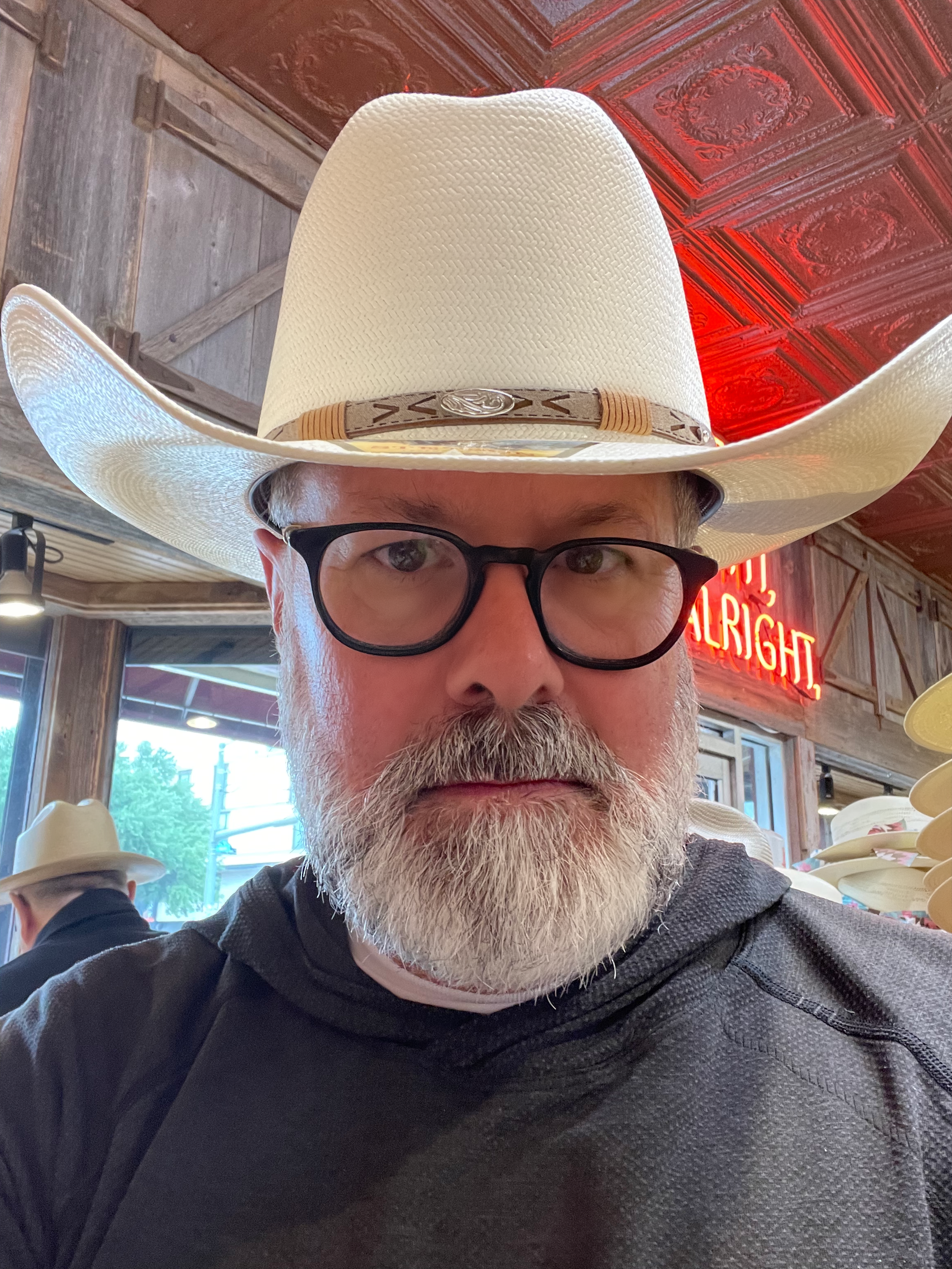 A man with glasses and a beard wearing a white cowboy hat, sitting in a restaurant with a wooden interior and red neon sign, other patrons visible in the background.