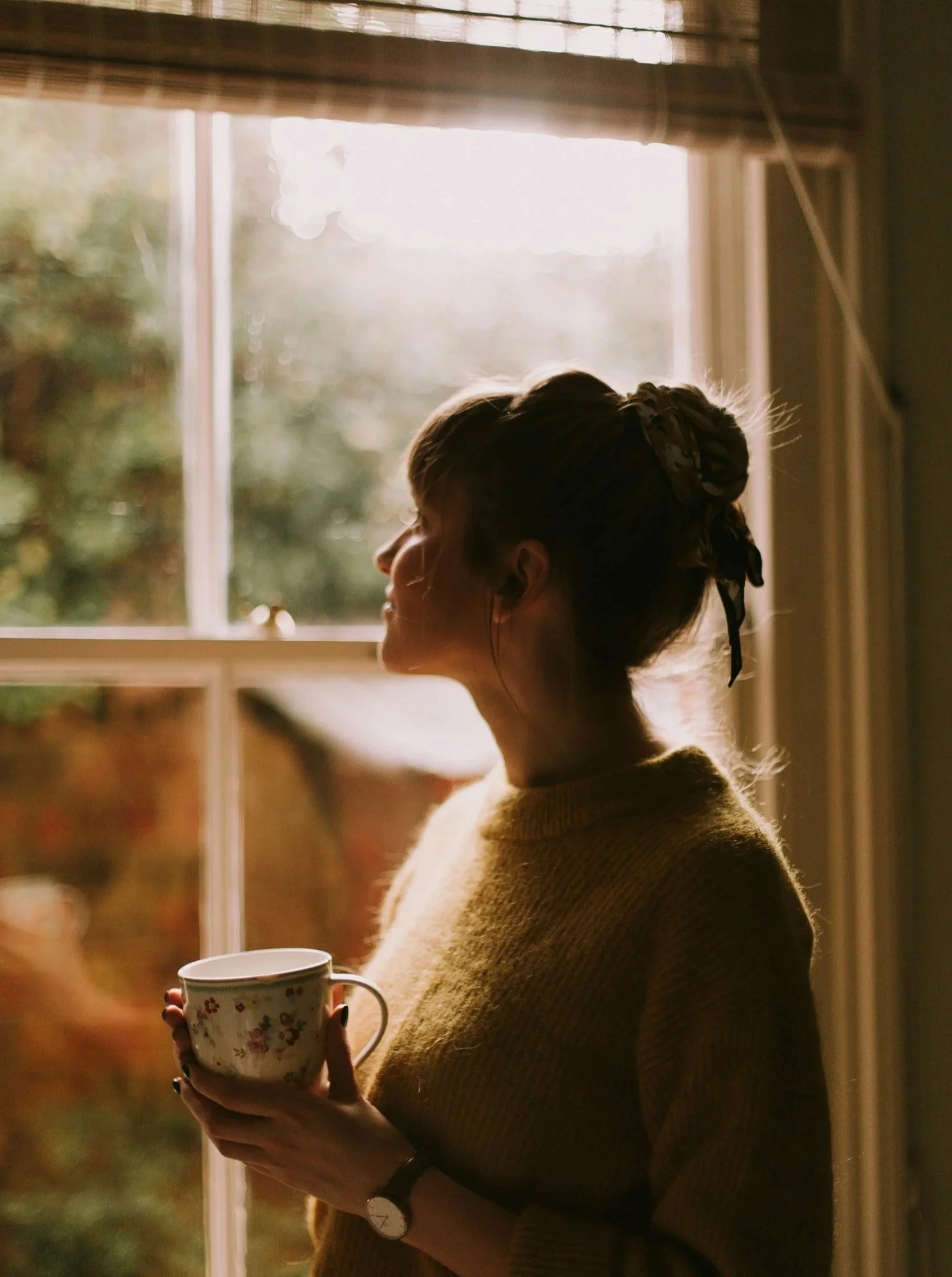 A woman standing by a window holding a floral mug, backlit by sunlight.
