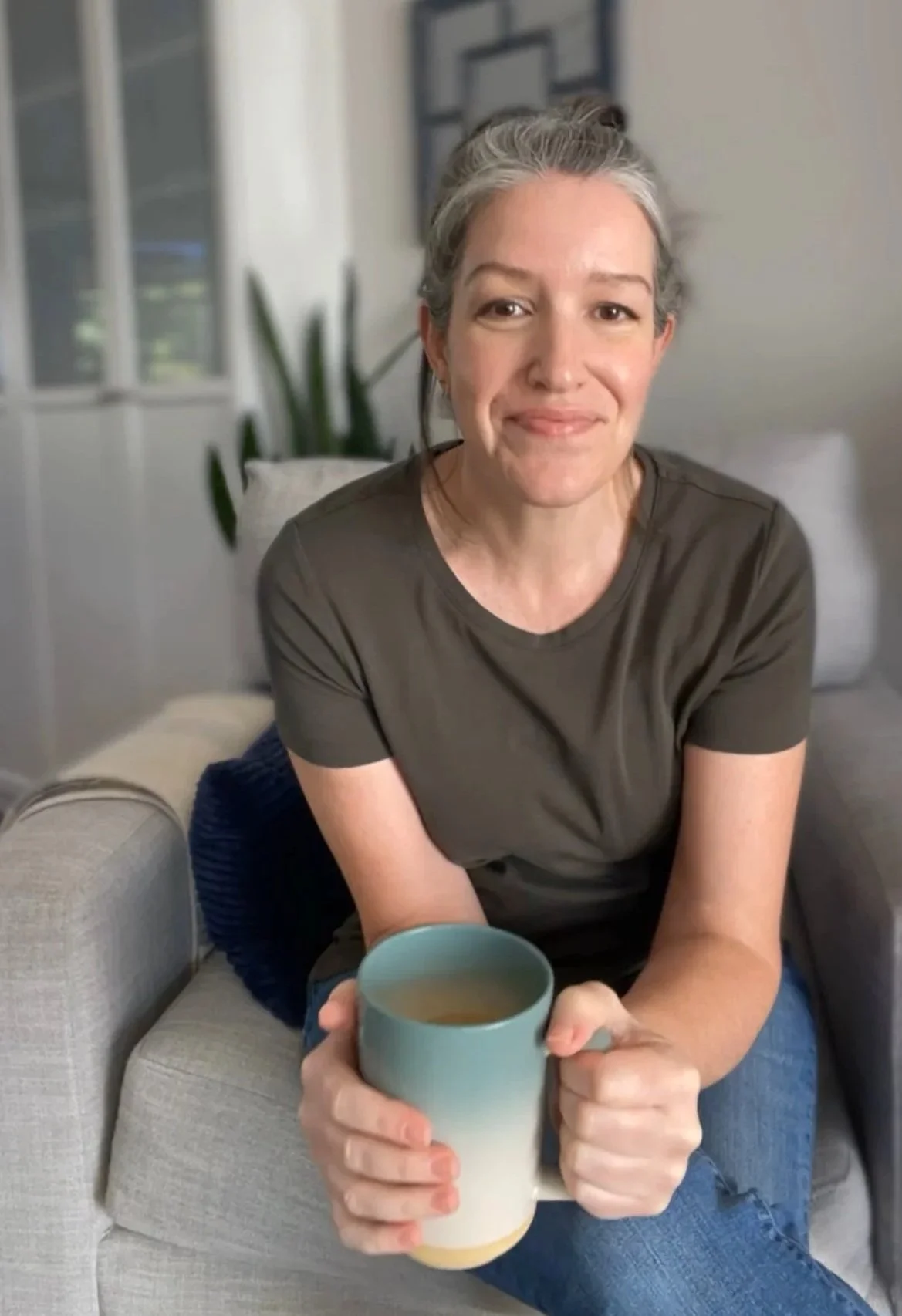 A photo of Michelle sitting on a couch, holding a steaming cup of coffee, smiling at the camera.