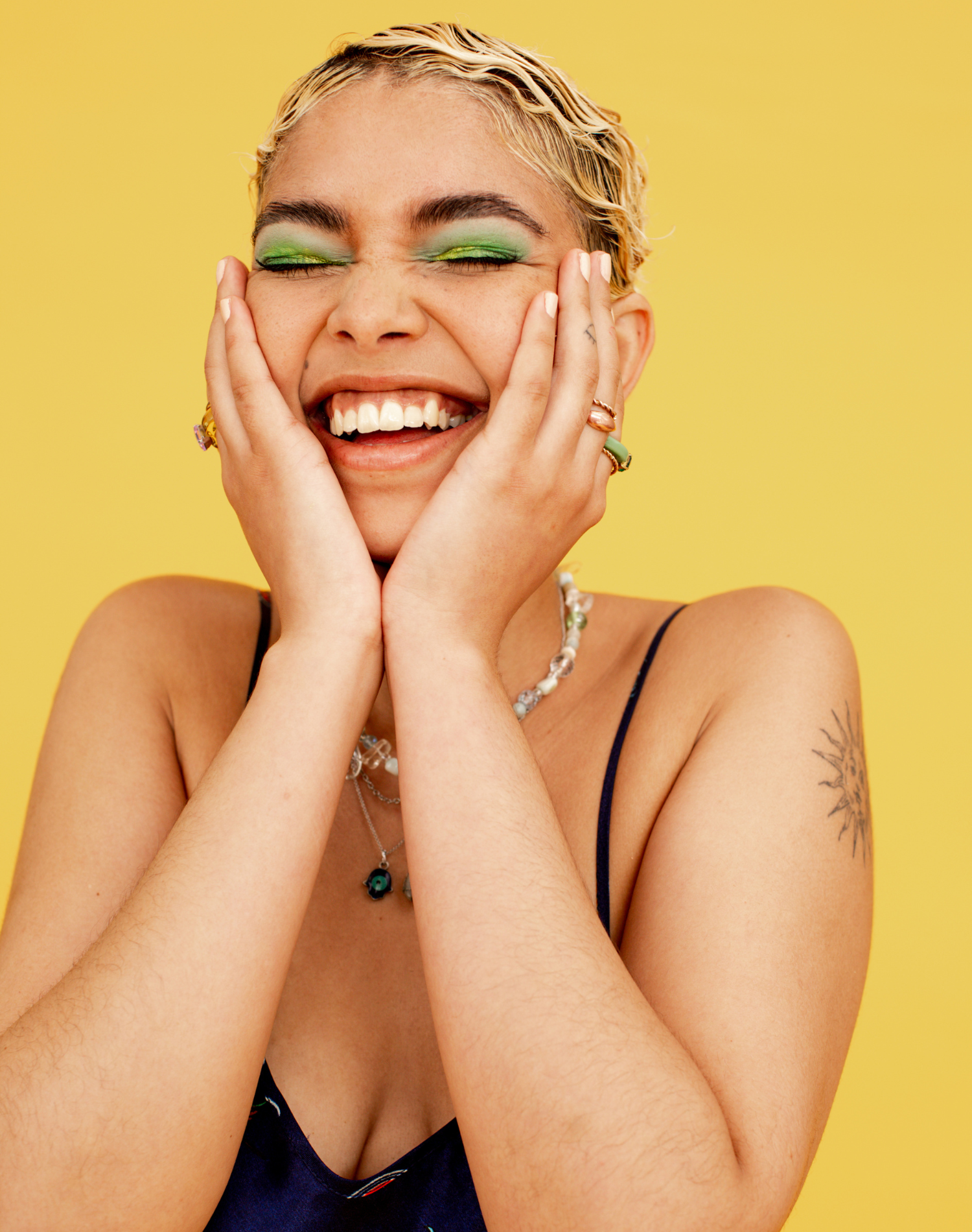 Woman with short blonde hair, green eye makeup, smiling with eyes closed, touching her face with hands, wearing jewelry, against a yellow background.