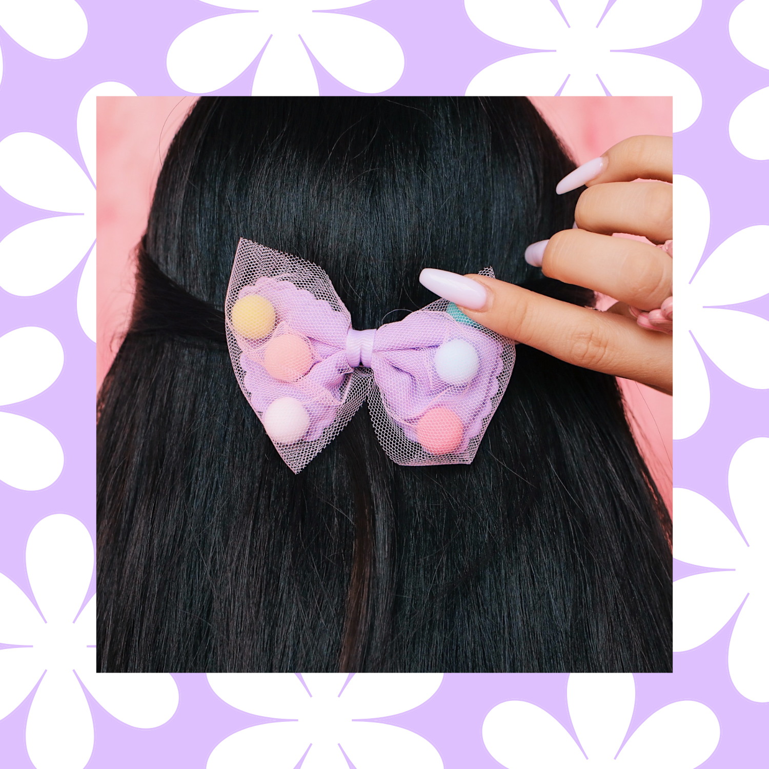 A person with long black hair is wearing a pastel pink and purple bow hair accessory decorated with pastel pom poms. Their hand with long white nails is touching the hair accessory.