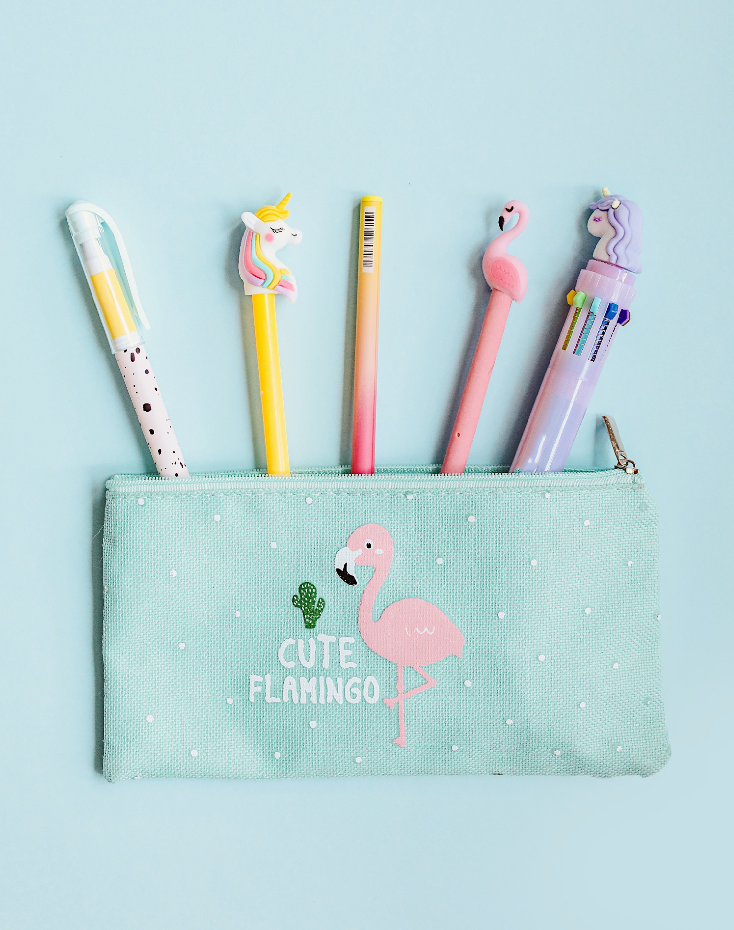 Set of colorful pens with unicorn and flamingo designs in a turquoise pouch with a pink flamingo and cactus illustration, on a light blue background.