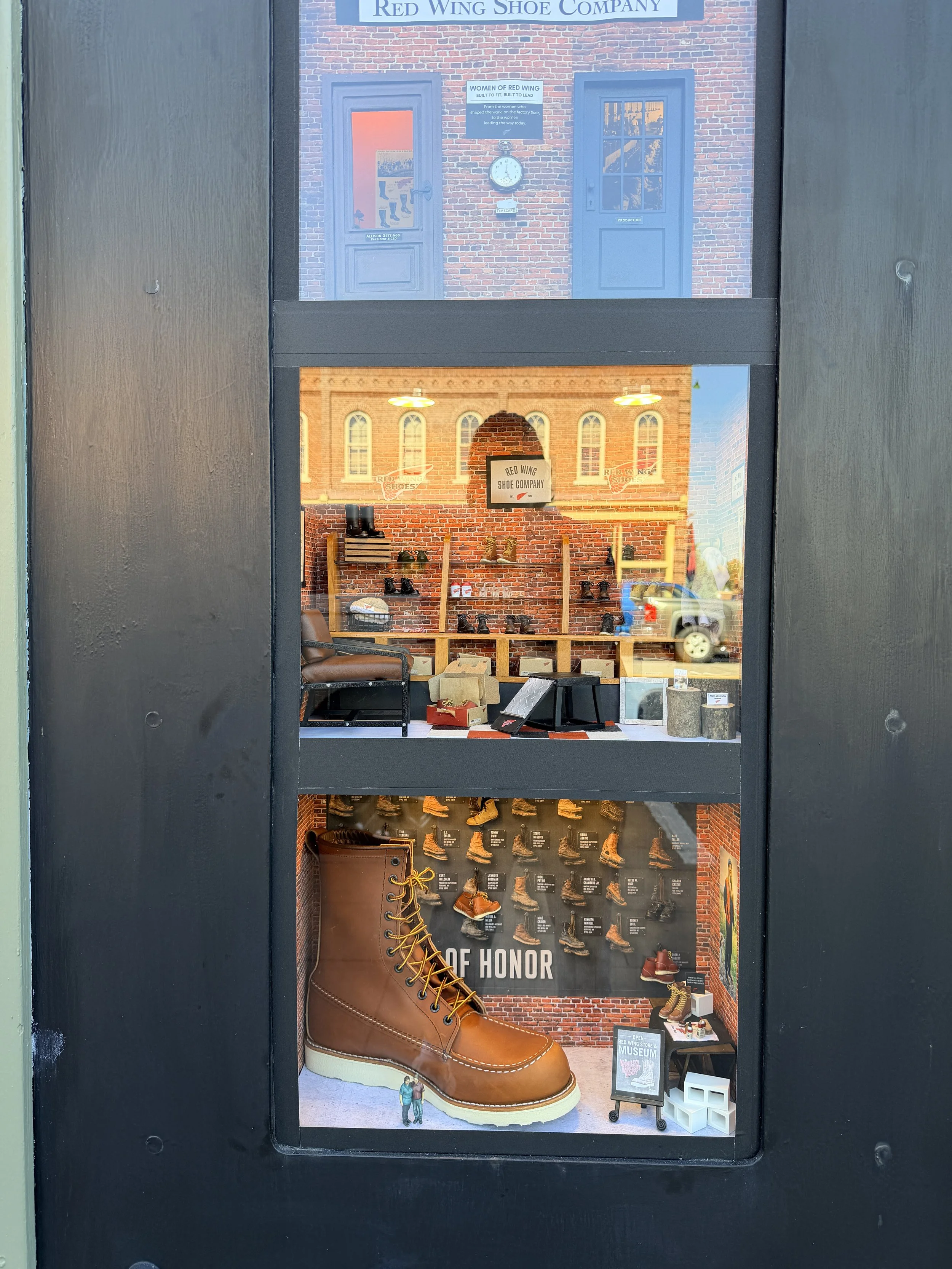 Display window of a shoe store featuring a giant brown leather work boot, shelves with various boots and shoes, and a brick wall background with signs and a logo for Red Wing Shoe Company.
