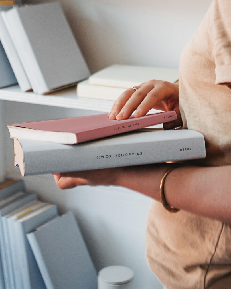 Person holding two books, one pink titled 'Hope in the Dark' and one gray titled 'New Collected Poems' in a room with bookshelves.