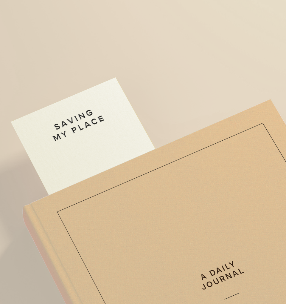 A closed tan-colored journal with a black rectangular border on the cover, titled "A Daily Journal." A white card labeled "Saving My Place" is inserted into the journal.