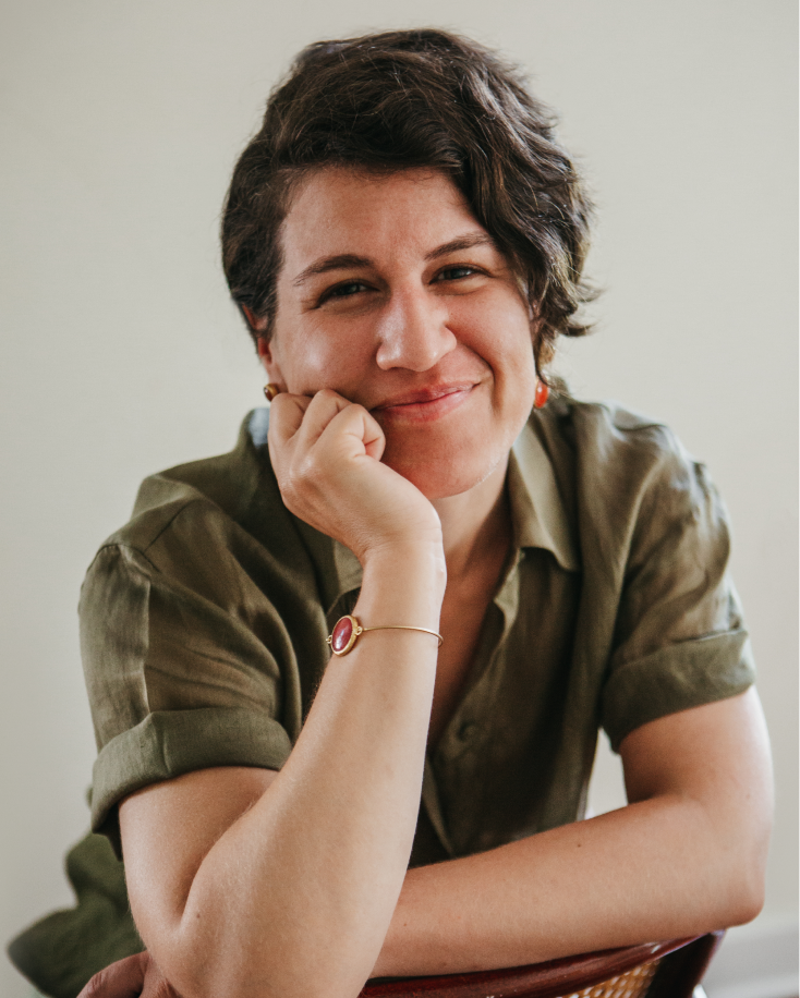 A woman with short, dark, wavy hair smiling gently, resting her chin on her right hand, wearing a green button-up shirt, a red bracelet, and small earrings, in front of a plain light background.