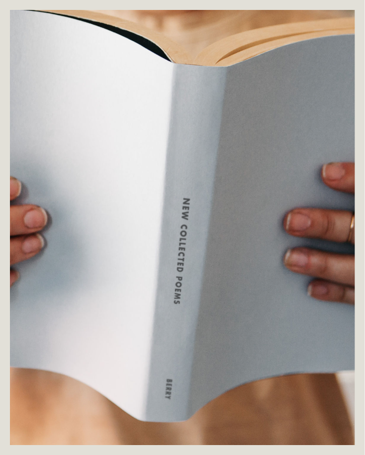 Person holding an open book with a blue cover titled 'New Collected Poems' by Wendell Berry, reading a book with visible pages.