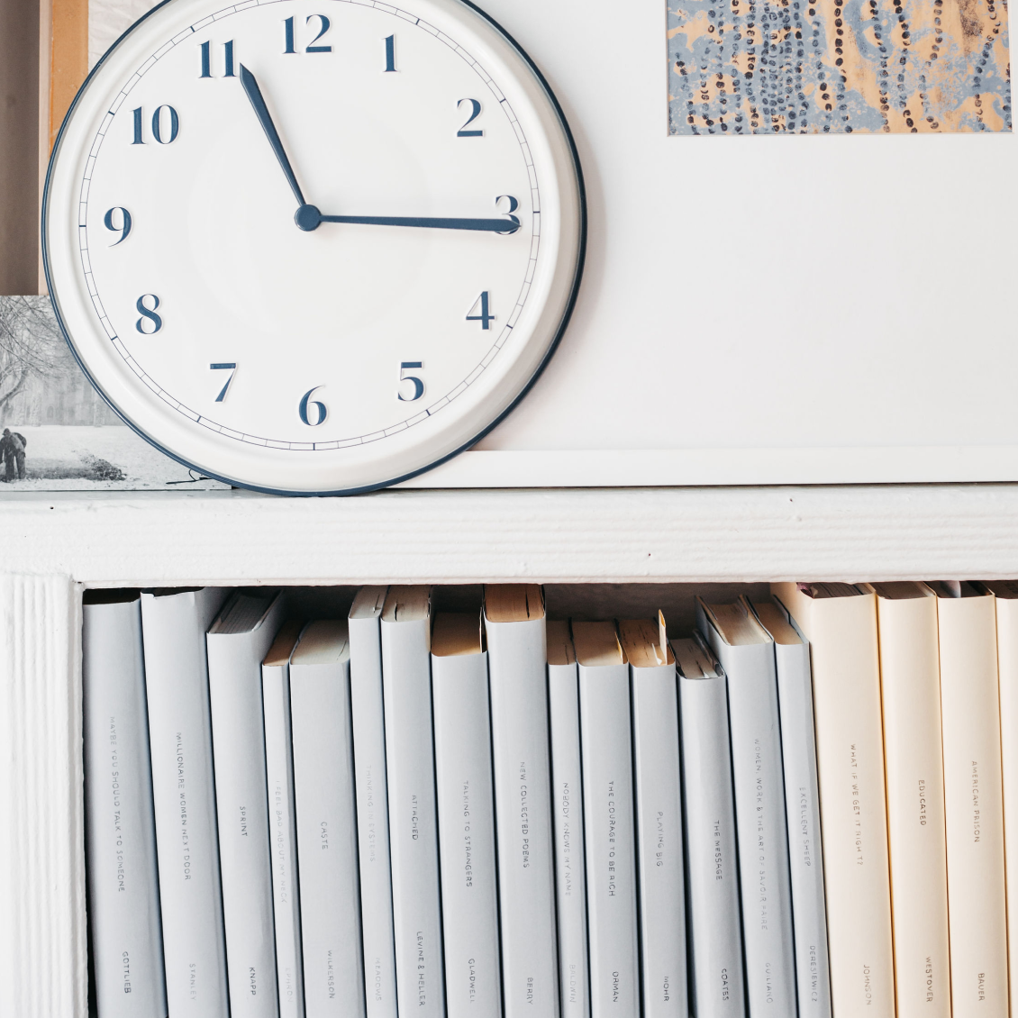 A row of white books in a modern home, titles facing outward. A mix of art in and black and white photography in the background.