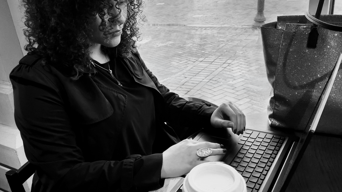 A black and white photo of P. Crismor working on her laptop, while seated at a cafe.