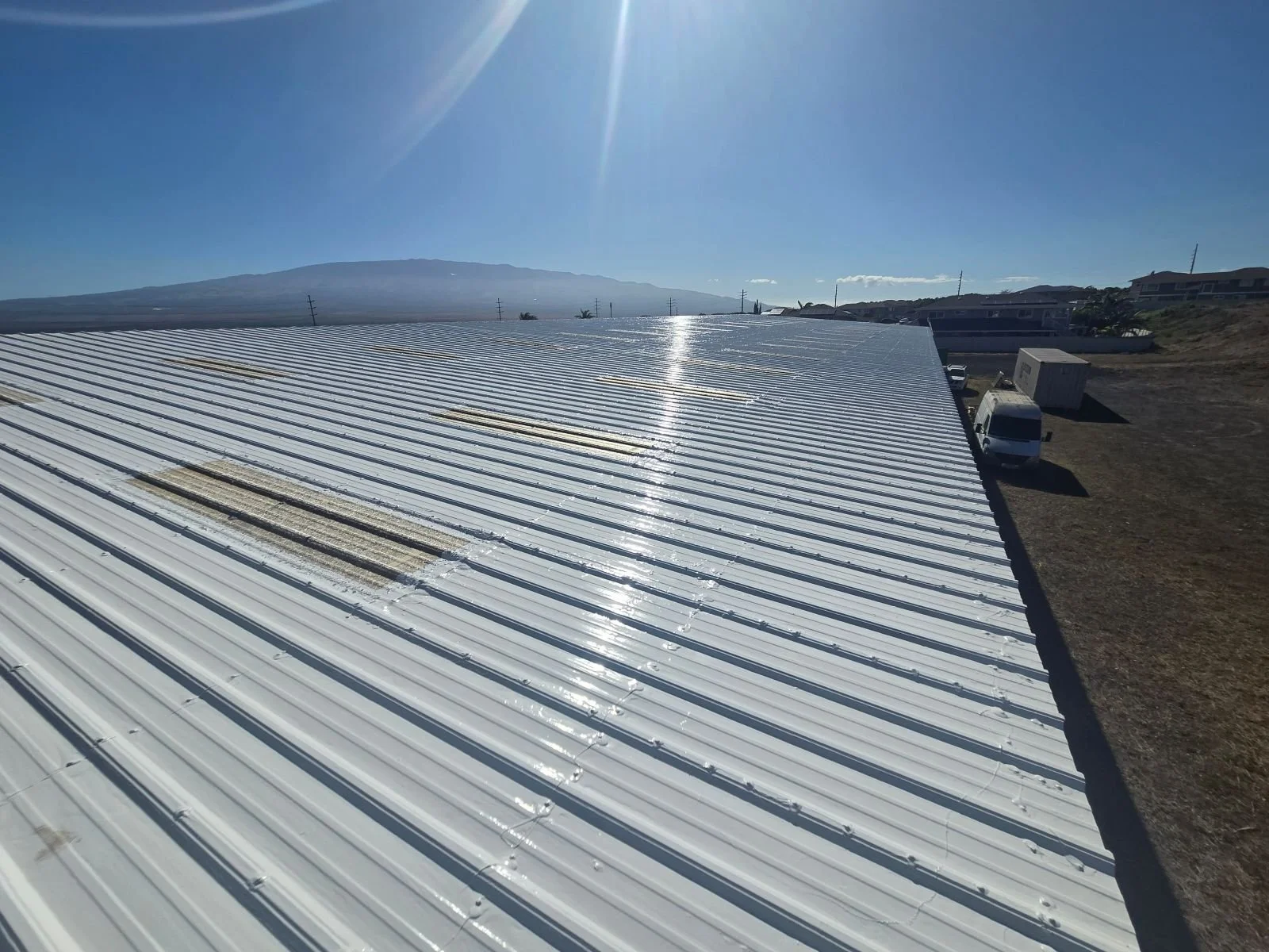 A wide view of a large, shiny, white metal rooftop with some rust-colored streaks and dirt, under a clear blue sky with the sun shining brightly.
