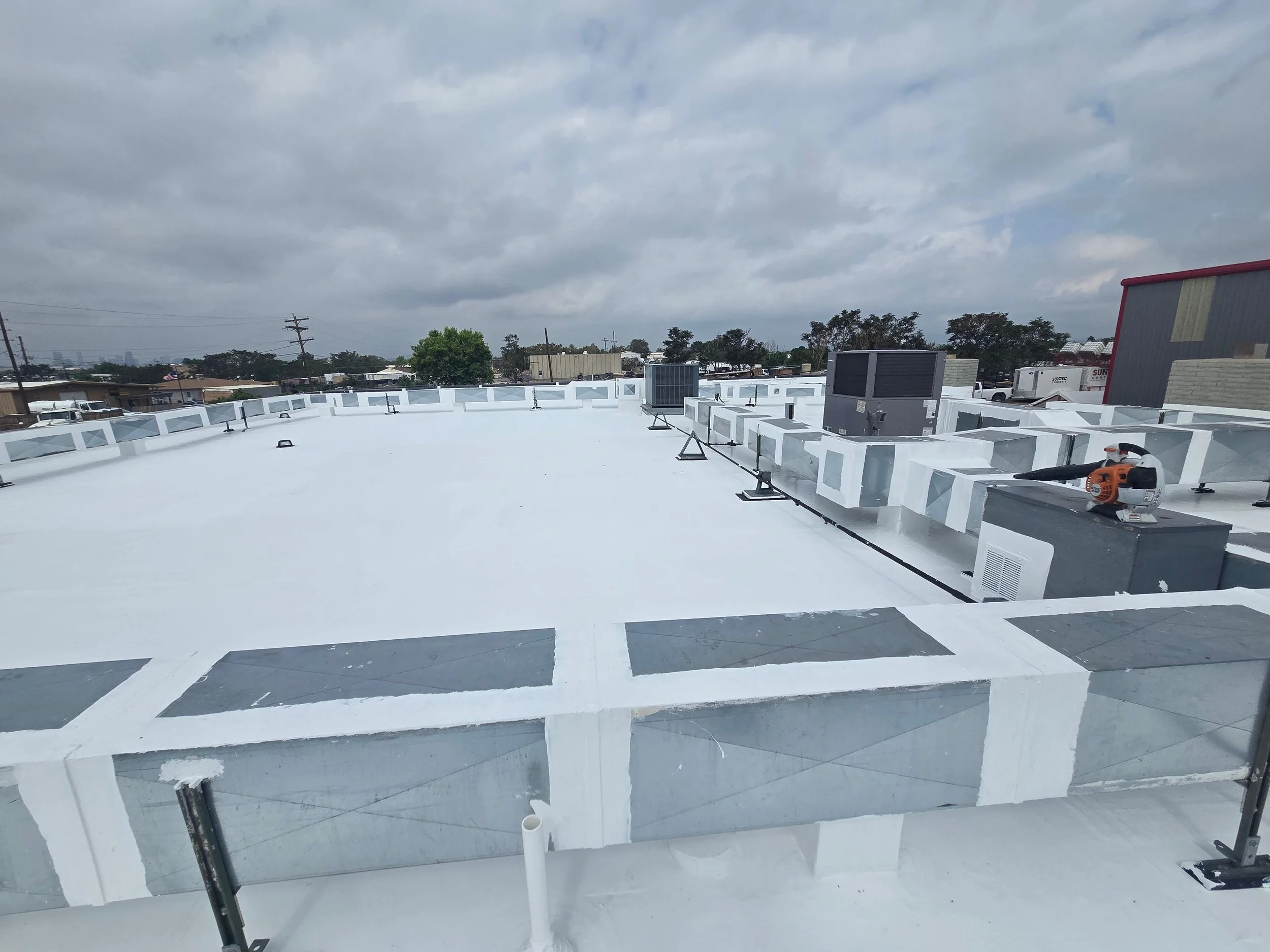 View from a rooftop under construction with a white membrane, HVAC units, tools, and neighboring buildings under a cloudy sky.