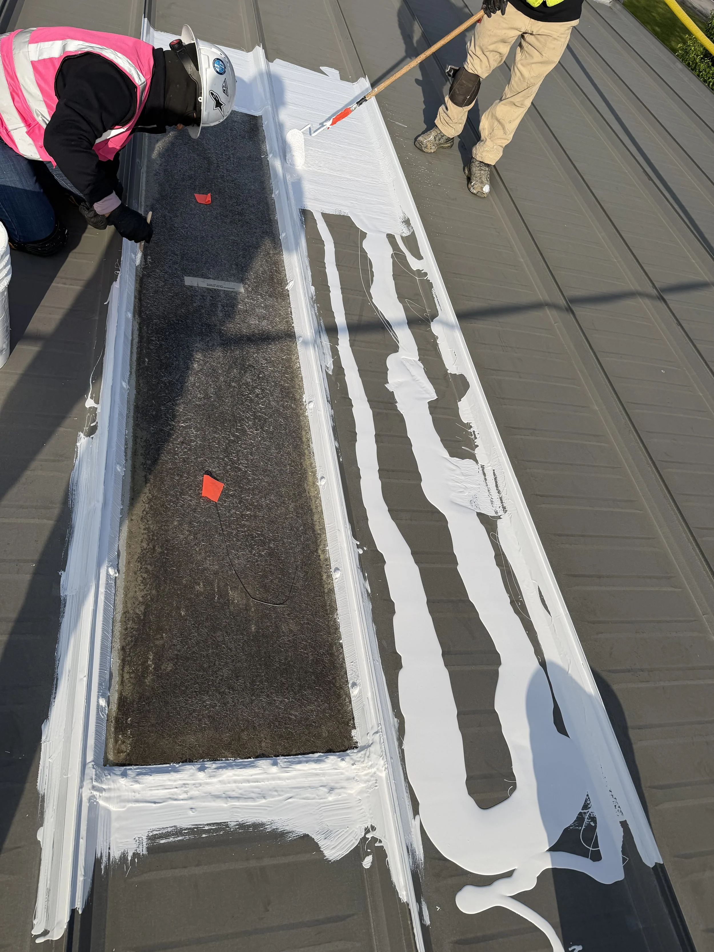 Two workers are applying white sealant around the edges of a rectangular window on a gray metal roof. One worker is kneeling, wearing a pink safety vest and a helmet, while the other is standing, using a roller brush.