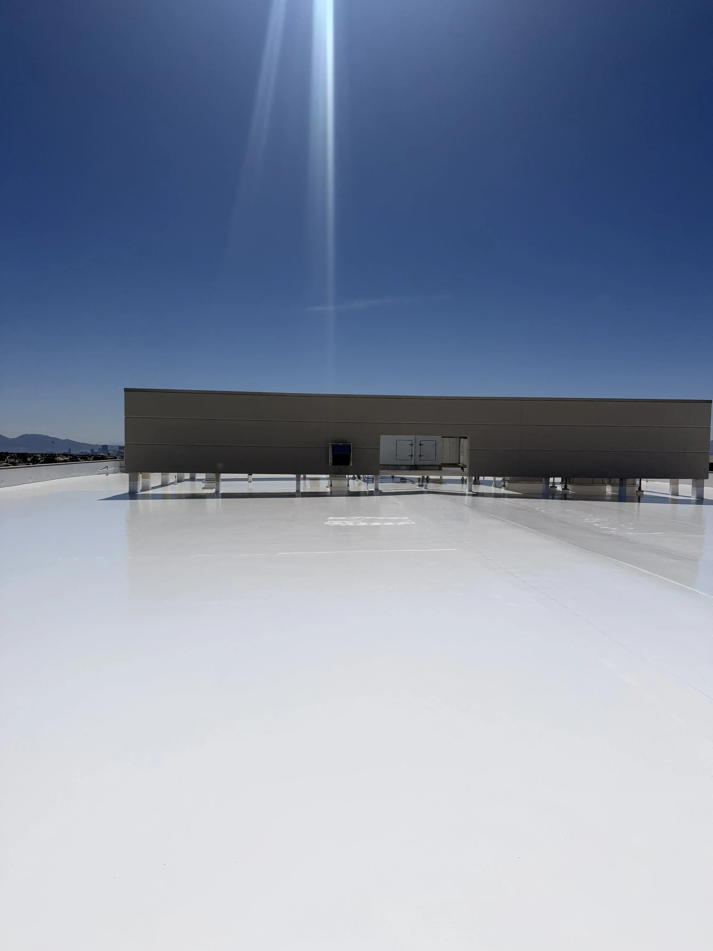 A large rooftop with a flat white surface, a rectangular building structure, and a bright blue sky with sunlight rays and a contrail.