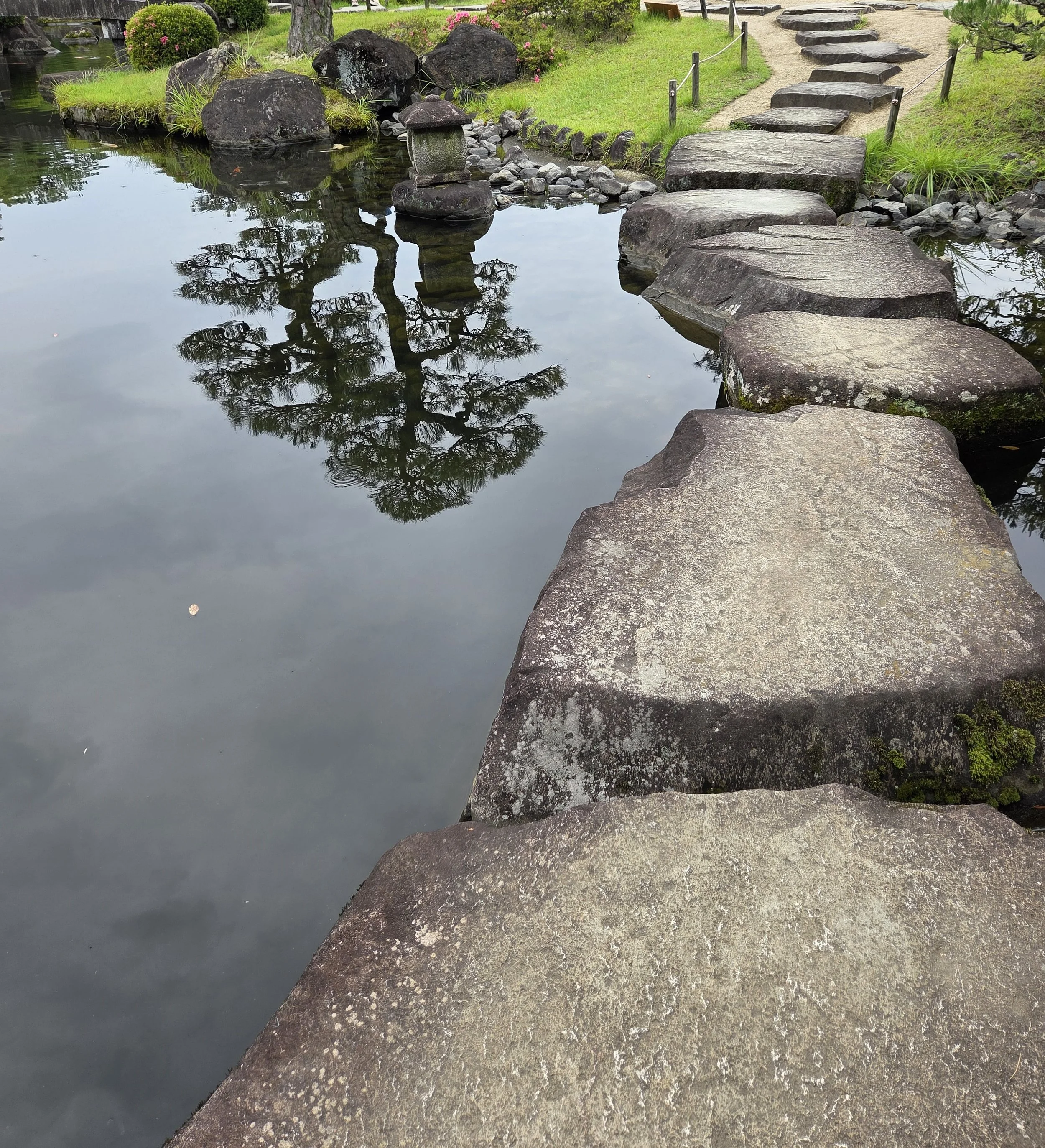 A stone pathway crossing a small pond in a Japanese garden, with rocks, greenery, and a reflection of a tree in the water.