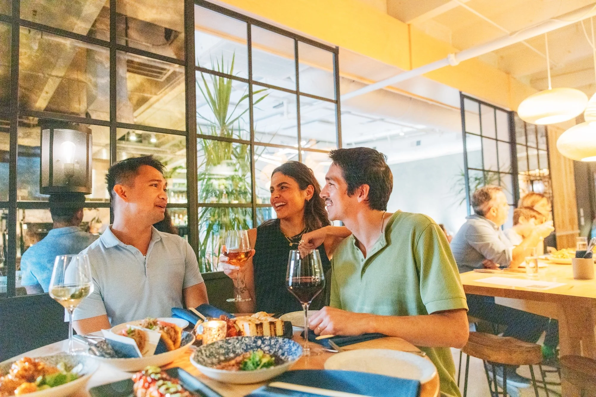 Three friends are sitting at a restaurant table, smiling and talking. They have drinks, including a glass of white wine and a glass of rosé, and there is a variety of food on the table. The restaurant has a modern, cozy atmosphere with warm lighting, large windows, and potted plants.