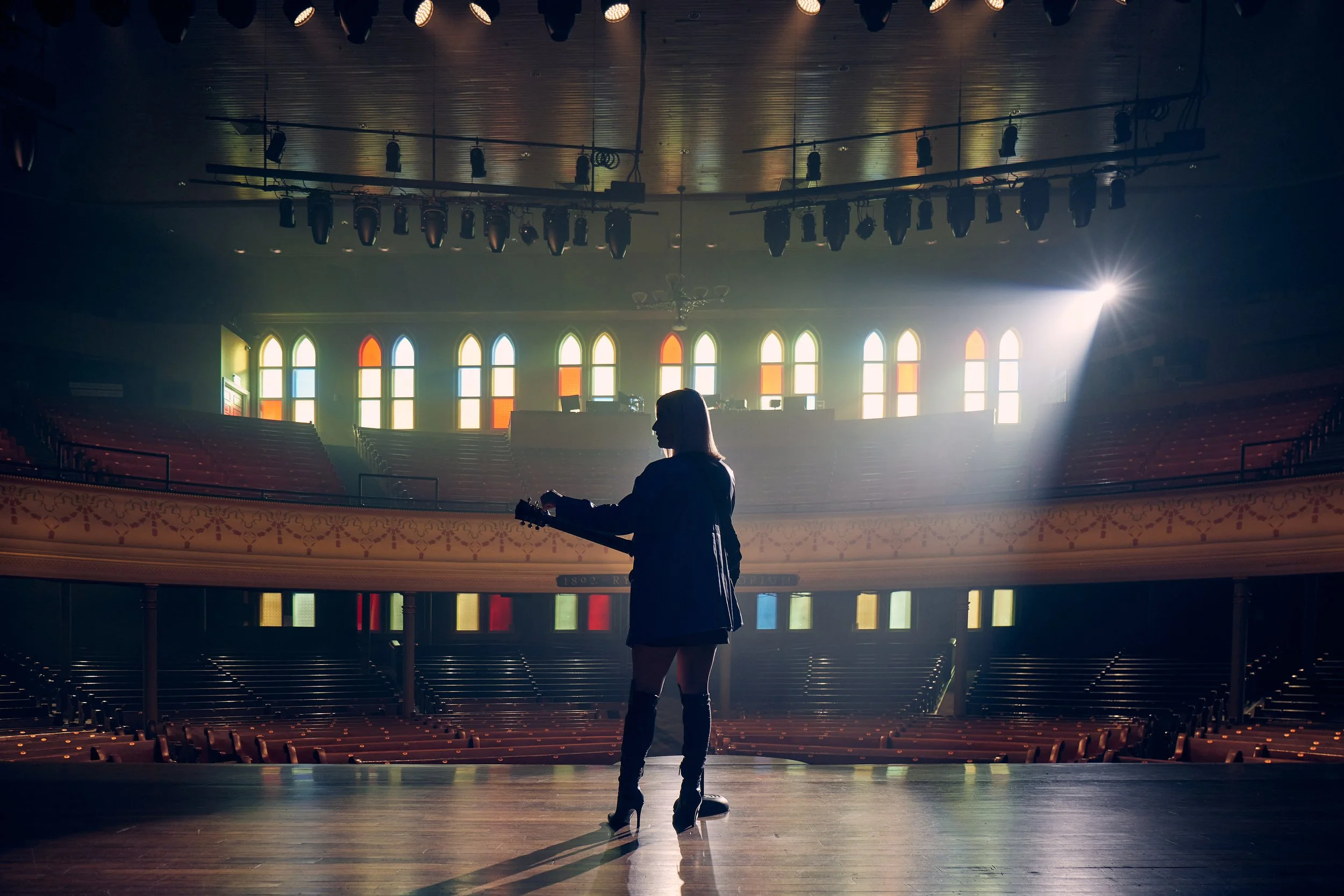 A musician standing on the Ryman stage with a guitar in a large, empty auditorium with colorful stained glass windows and stage lights.