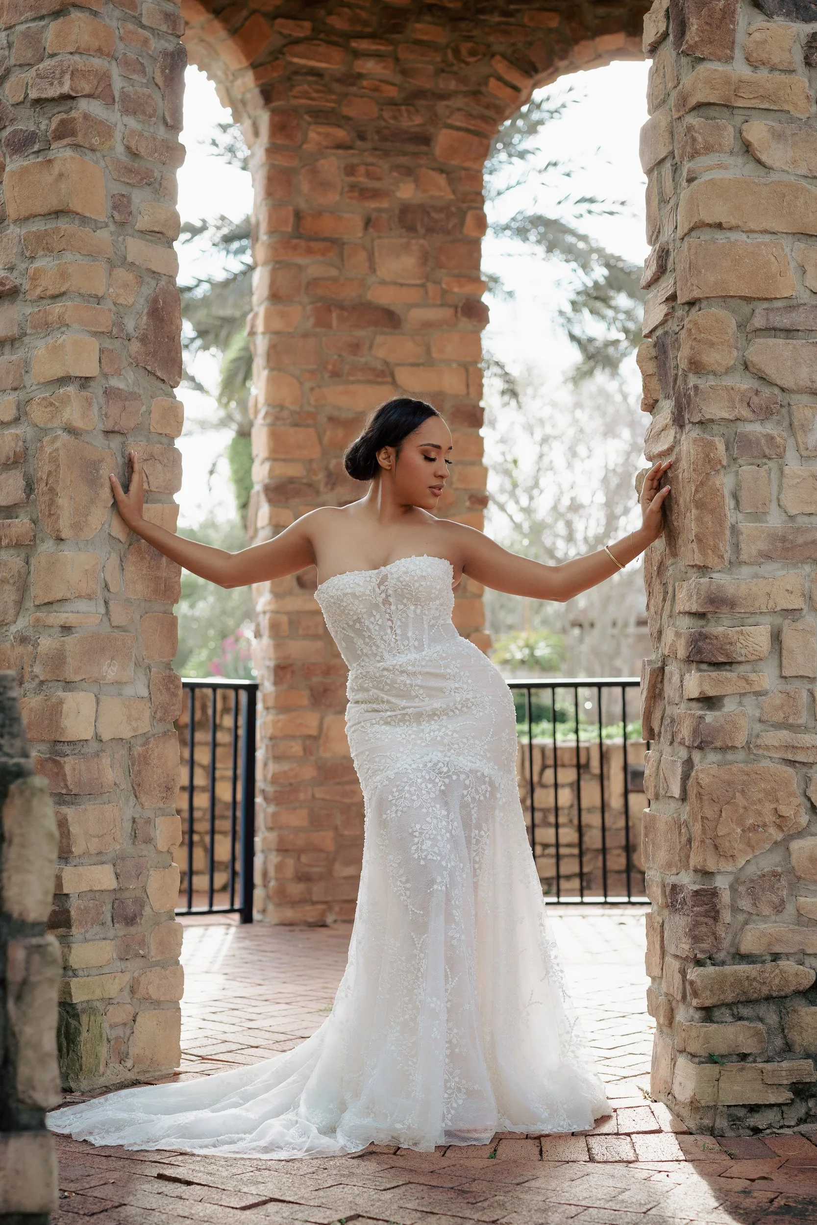 A woman in a white wedding dress with lace details stands between two brick pillars under a stone archway. She is looking down with her hands touching the pillars on either side. In the background, there are trees and a black metal fence.