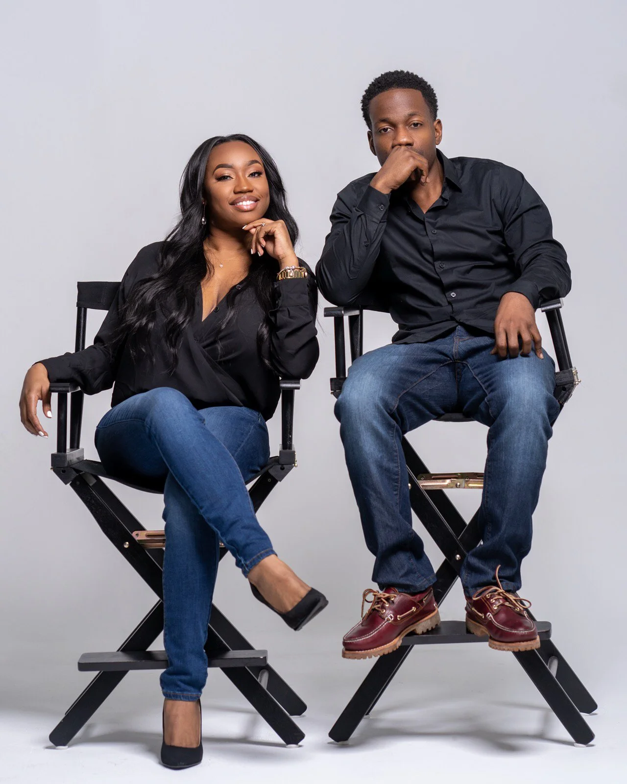 A man and woman sitting on director's chairs against a plain white background, both dressed in black shirts and blue jeans, posing confidently.