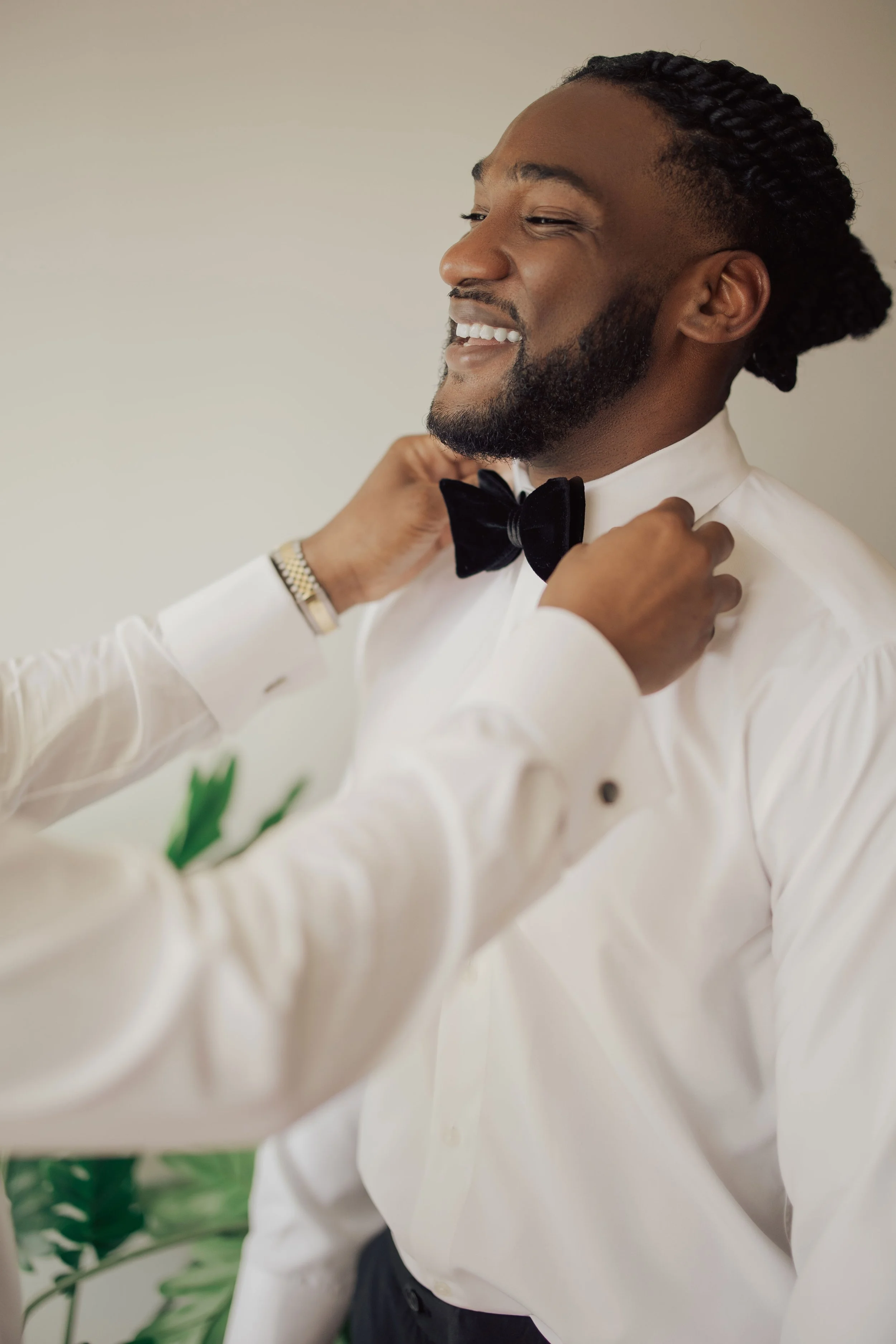 A man with a beard and braids smiling as someone adjusts his black bow tie, wearing a white tuxedo shirt, with greenery in the foreground.