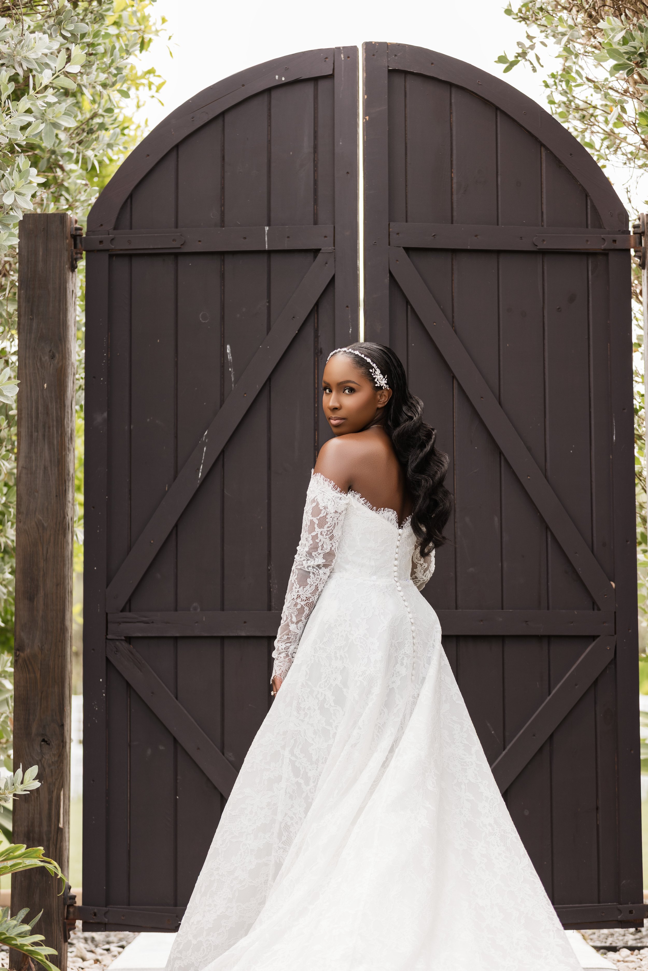A woman in a white wedding dress with lace long sleeves and floral headband standing in front of a large black wooden gate with greenery on the sides.