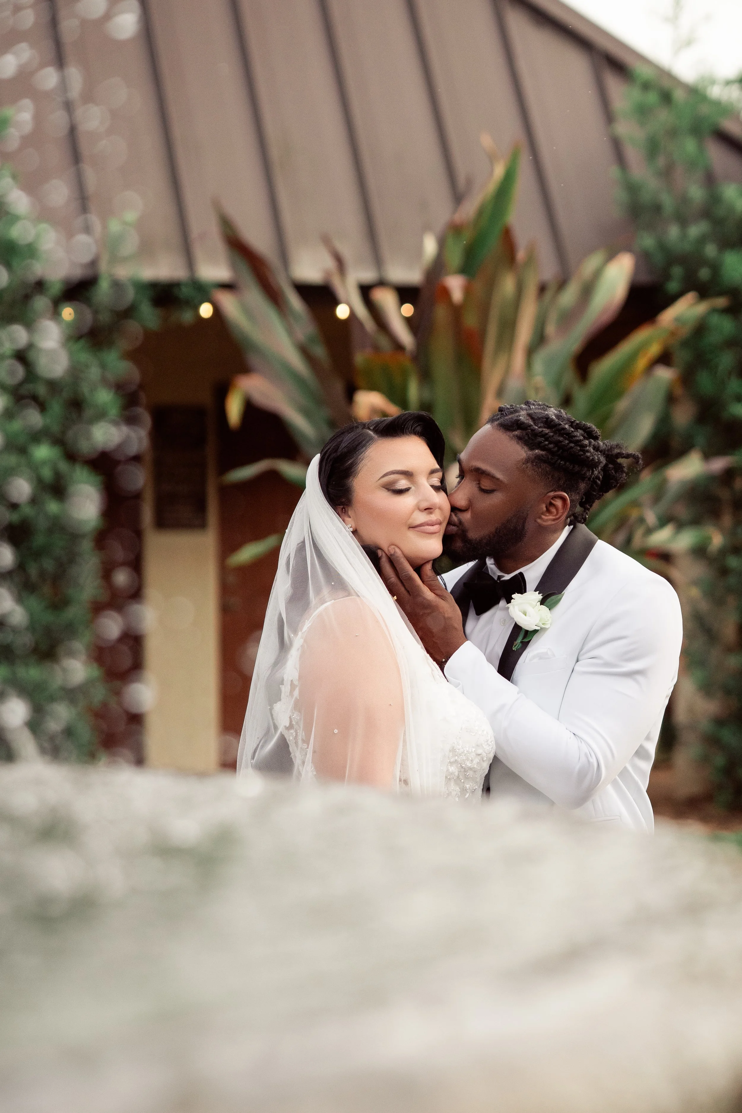 A bride and groom sharing a kiss during their wedding ceremony outdoors, with greenery and a wooden structure in the background.