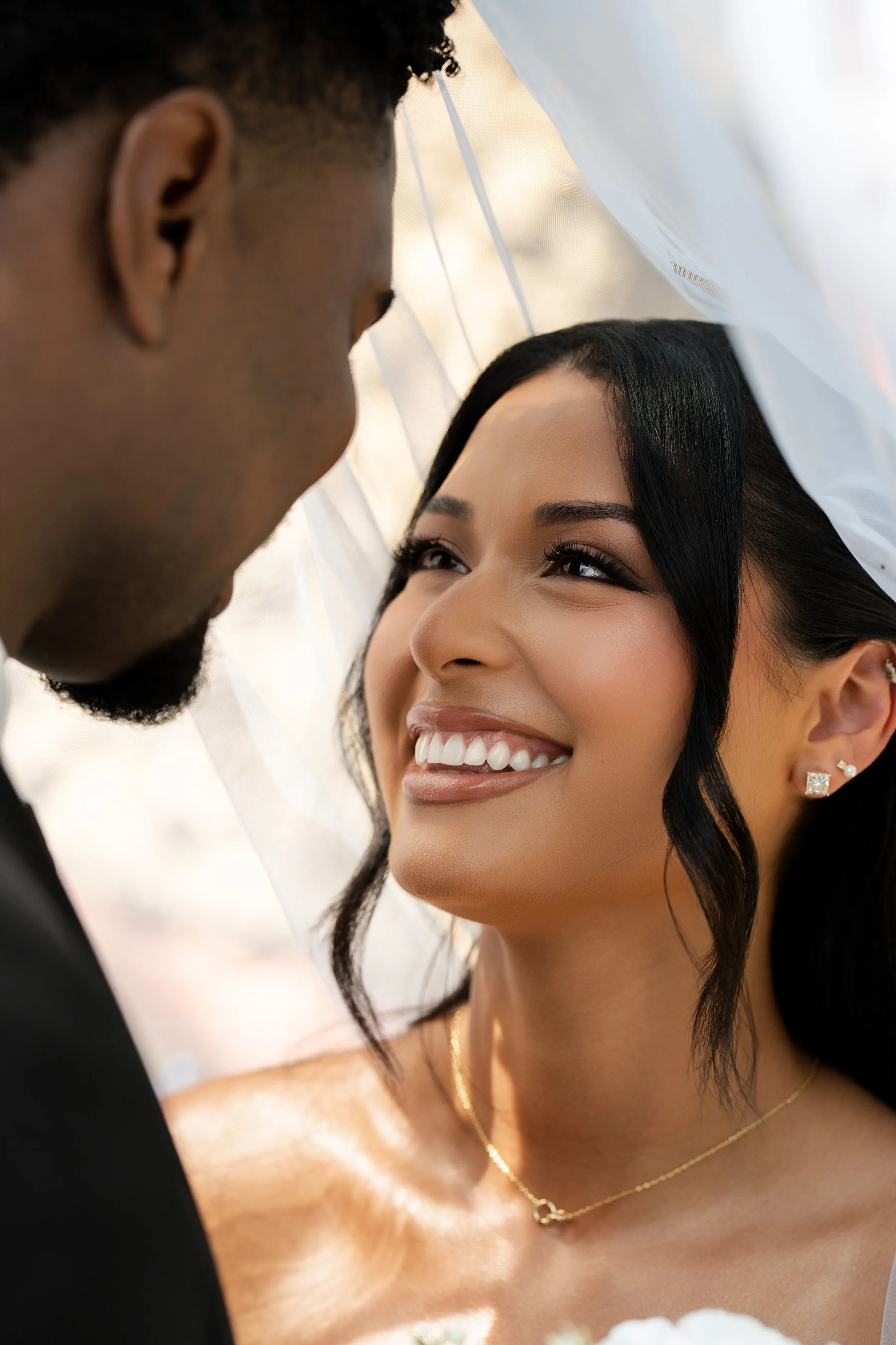 Close-up of a smiling woman with black hair and earrings, with a man near her, possibly in a wedding setting.